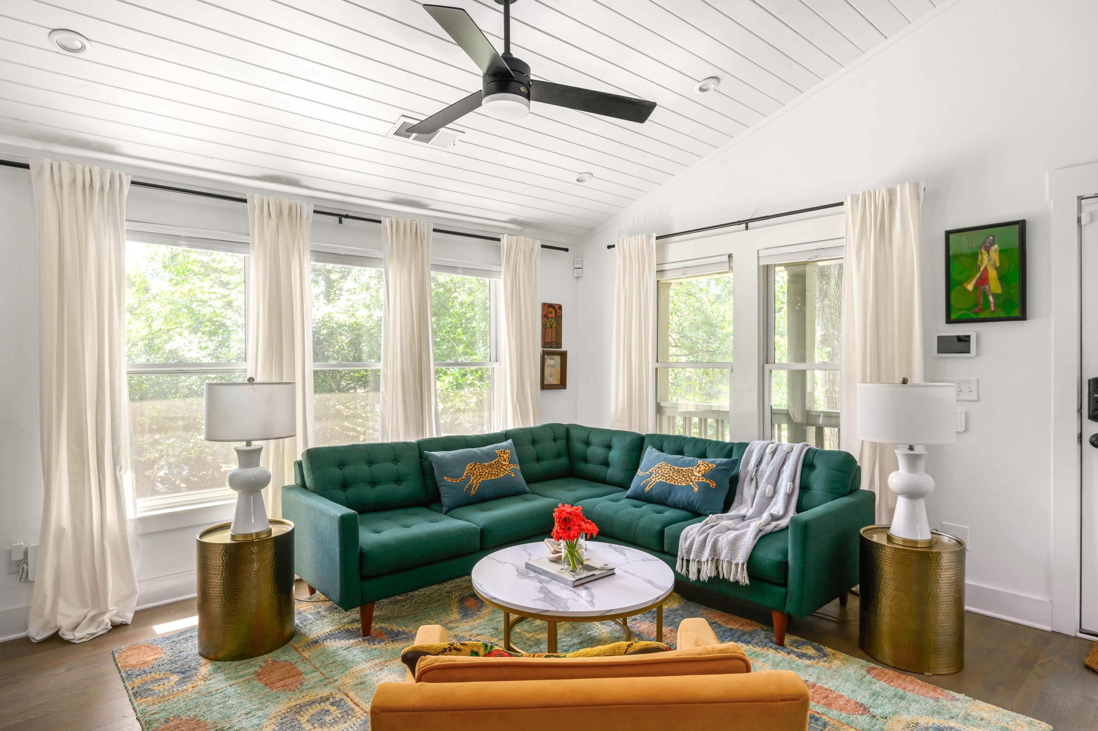 The living room features a green upholstered sectional sofa, a round coffee table with a floral arrangement, and large windows draped with light-colored curtains.