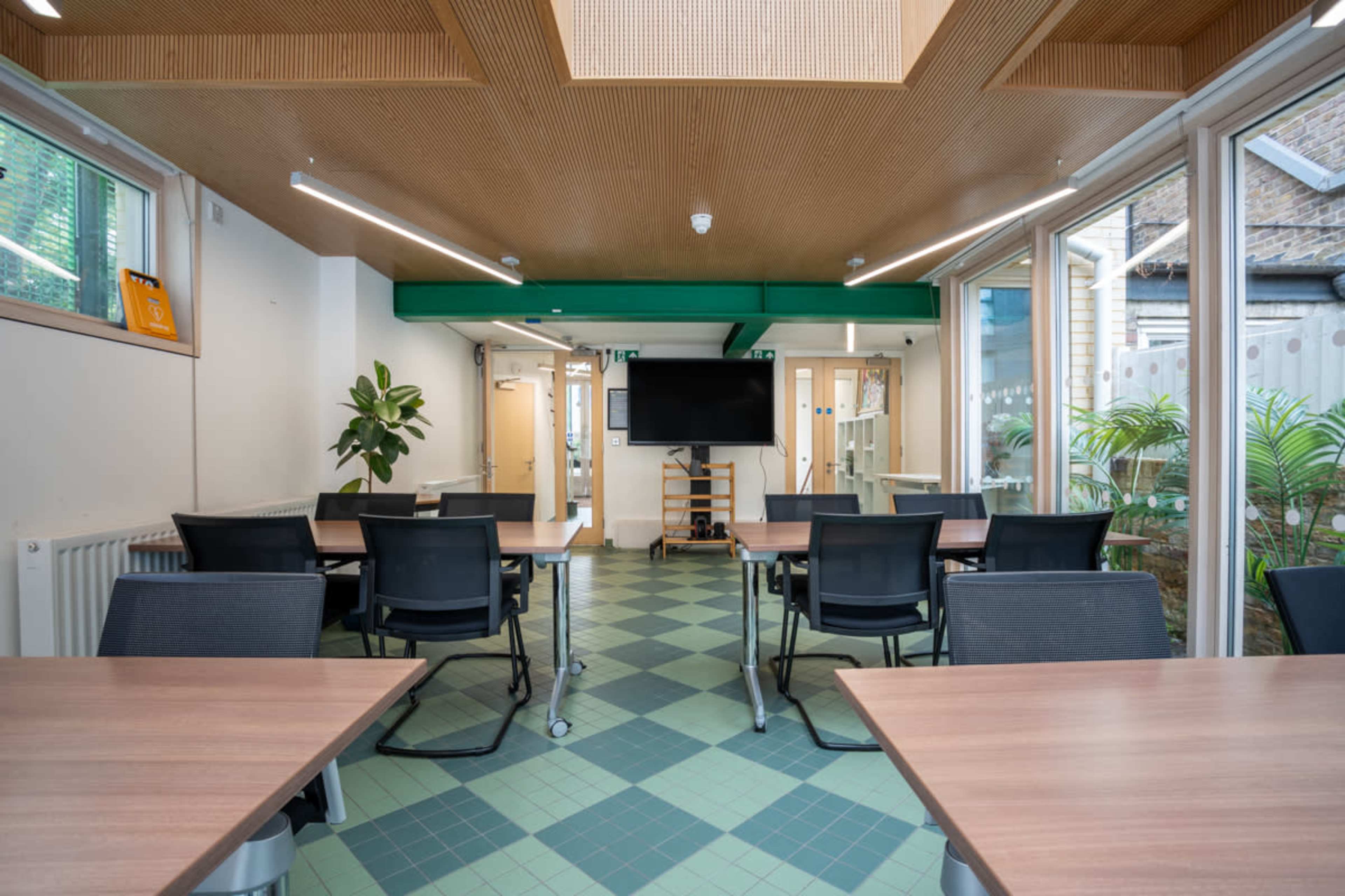 A modern classroom with several tables and chairs arranged around a central area, featuring large windows and a wall-mounted screen.