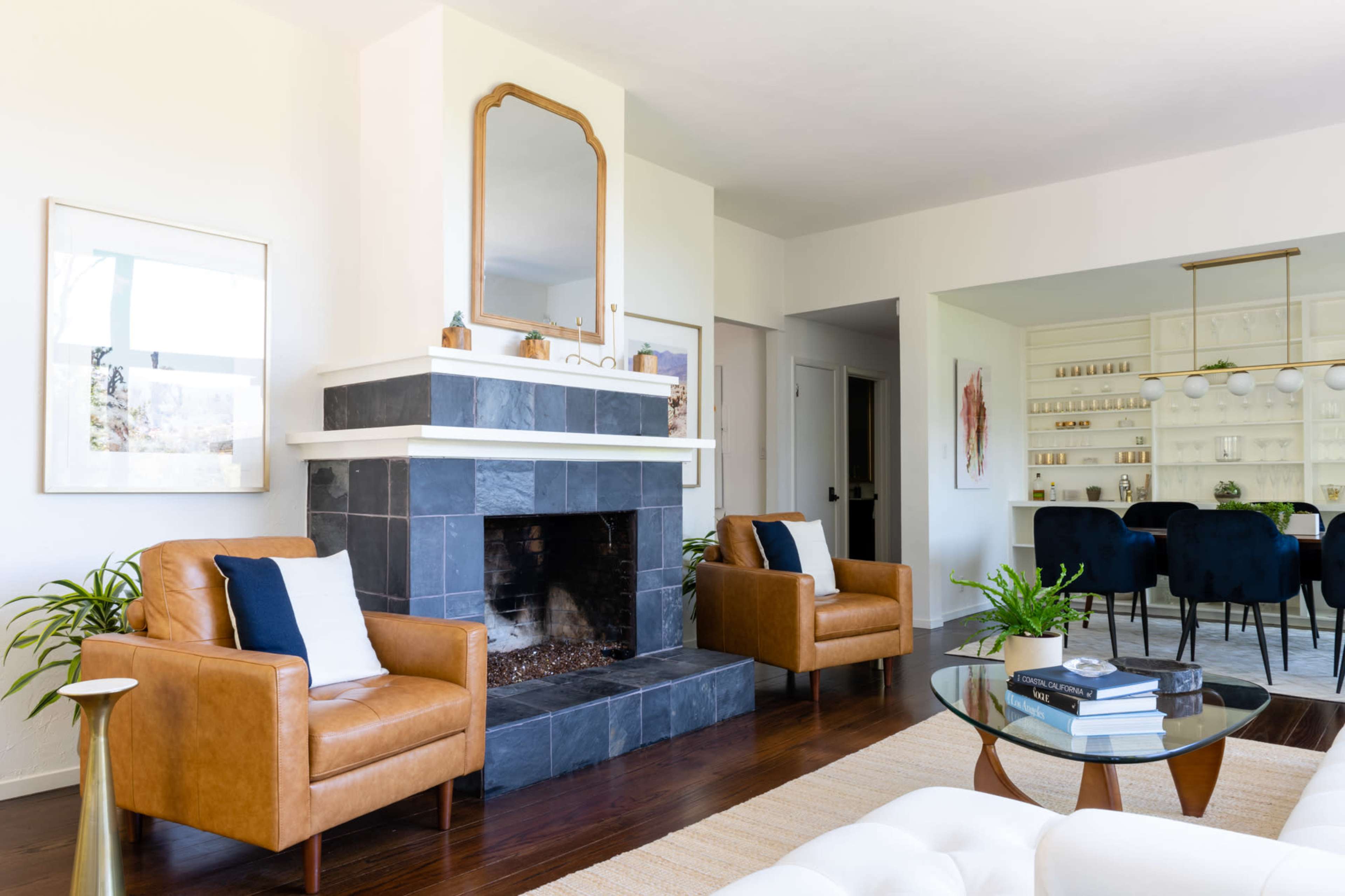 A modern living room features a slate fireplace, two brown leather chairs, a glass coffee table, and an adjacent dining area with a white cabinet and blue chairs.
