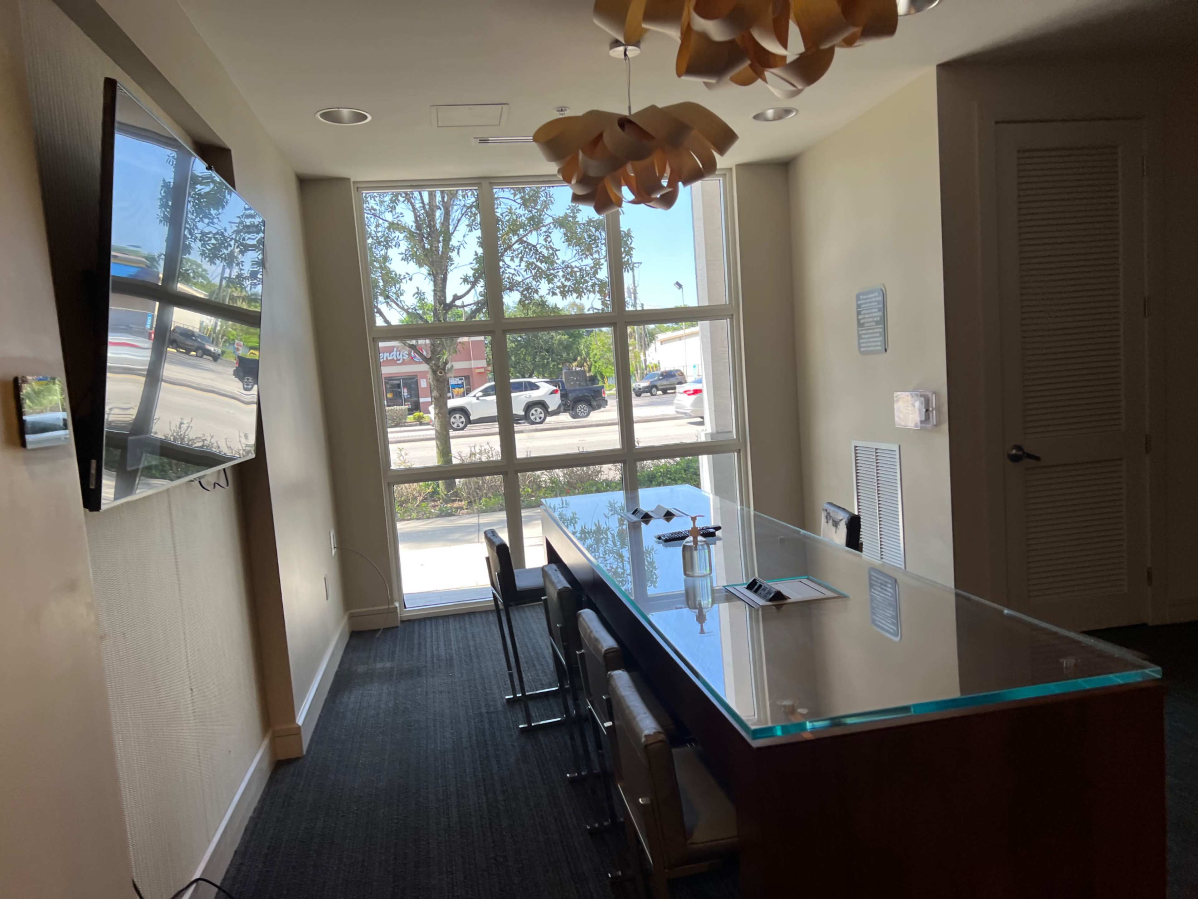 A meeting room features a long glass table, chairs, a television on the wall, and large windows providing a view of the street outside.