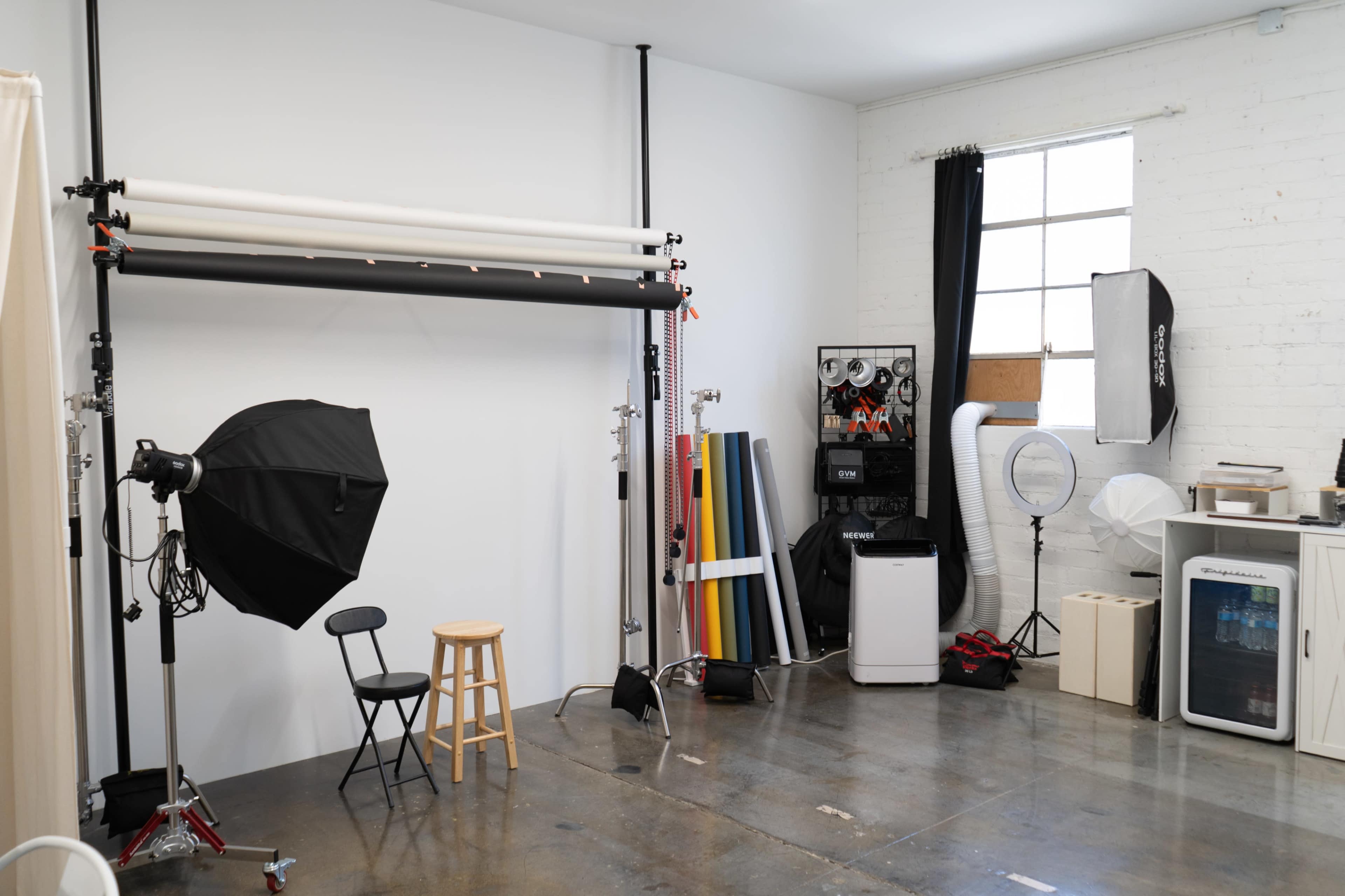 The image shows a photography studio with various lighting equipment, backdrops, and a chair arranged in a spacious, bright room.