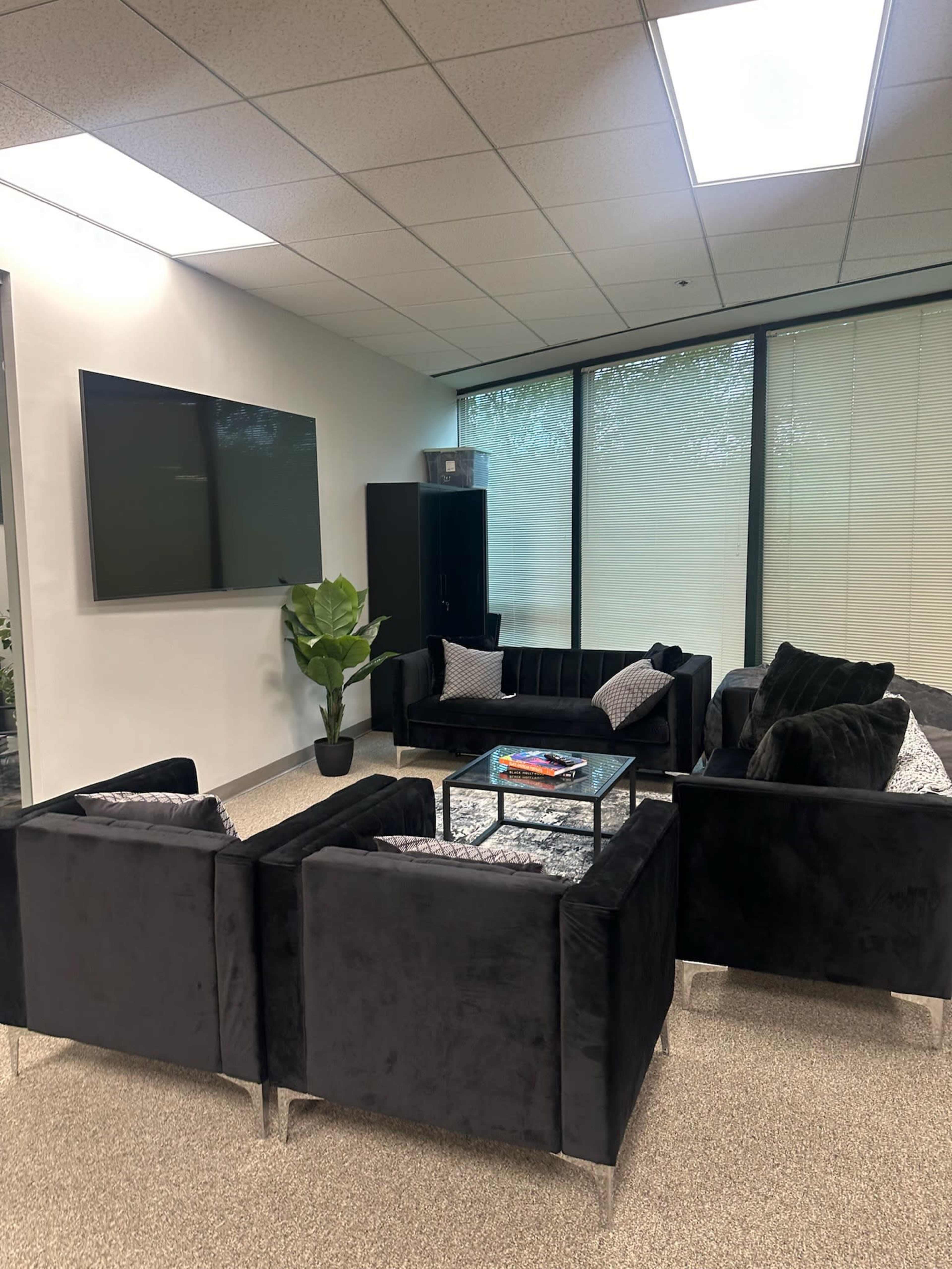The image shows a modern office waiting area featuring dark, plush seating arranged around a glass coffee table, with a television mounted on the wall and large windows allowing natural light.