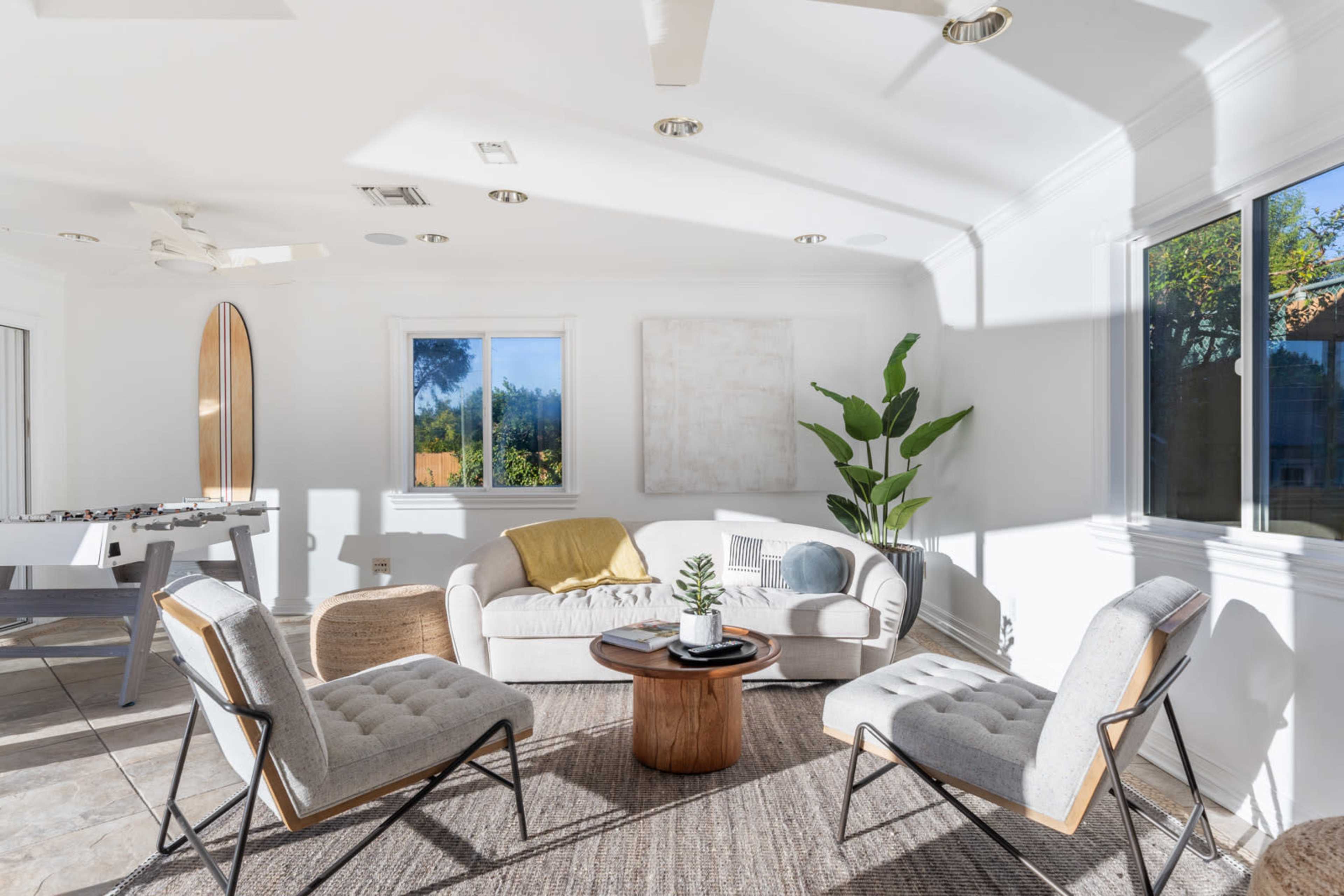 A bright, modern living room features a white sofa, two gray armchairs, a wooden coffee table, and a potted plant, with large windows allowing natural light to fill the space.