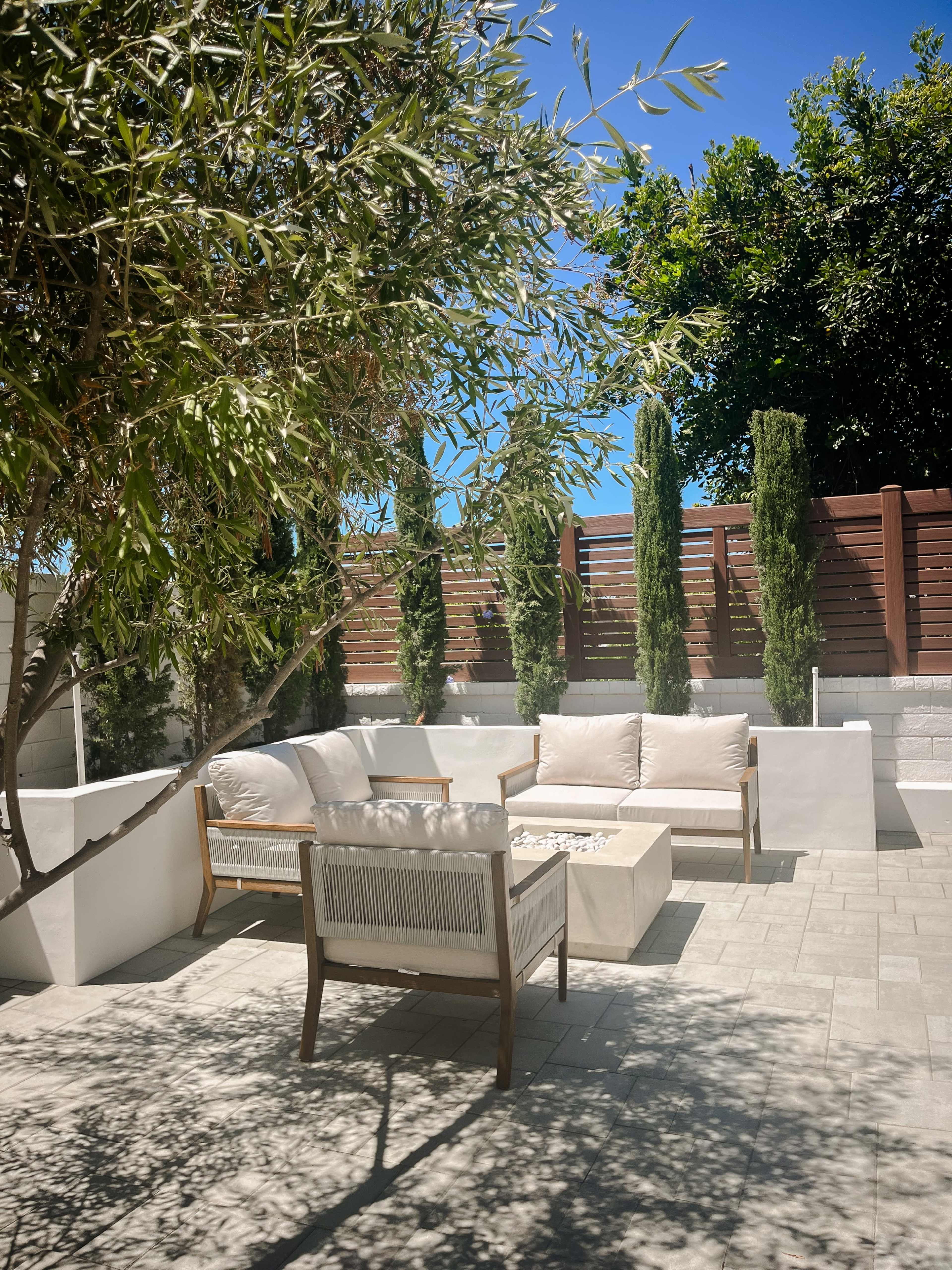 A modern outdoor seating area features beige couches and a coffee table surrounded by tall greenery and a wooden fence.
