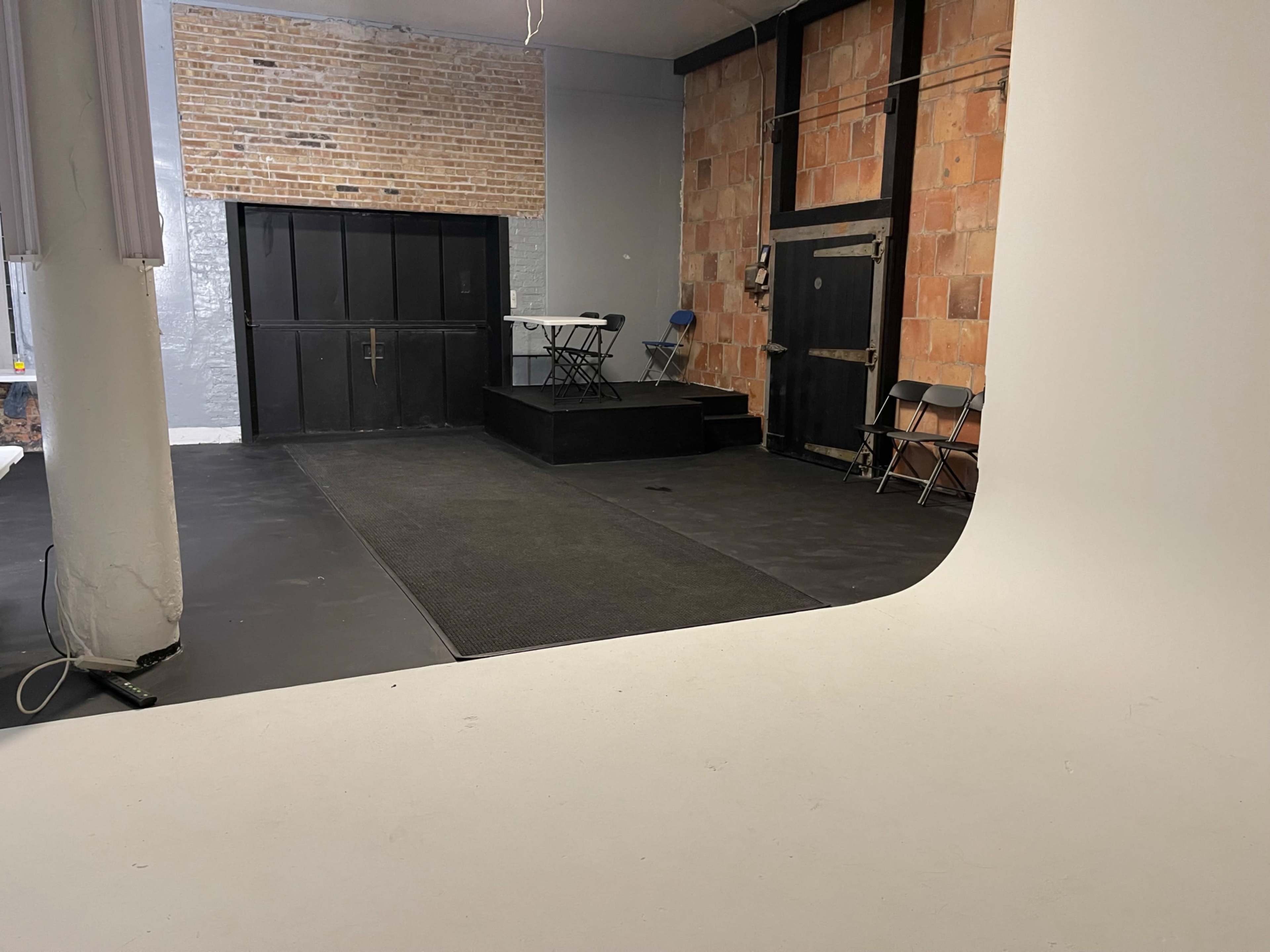 The image shows a spacious room with a black floor, a raised platform, and a brick wall, featuring a back door and a few metal chairs arranged nearby.