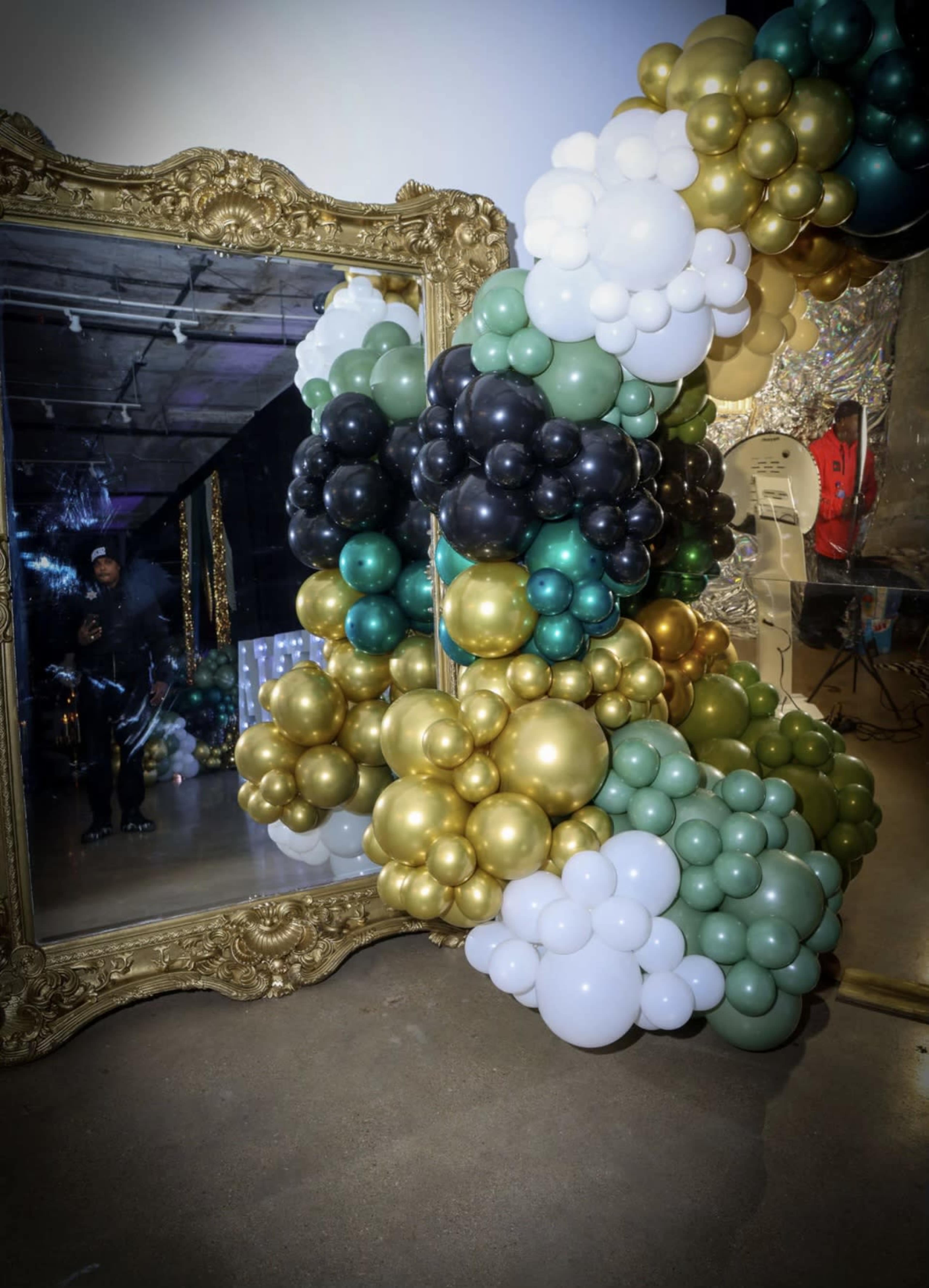 A large arrangement of colorful balloons in black, gold, green, and white surrounds an ornate mirror in a decorated space.