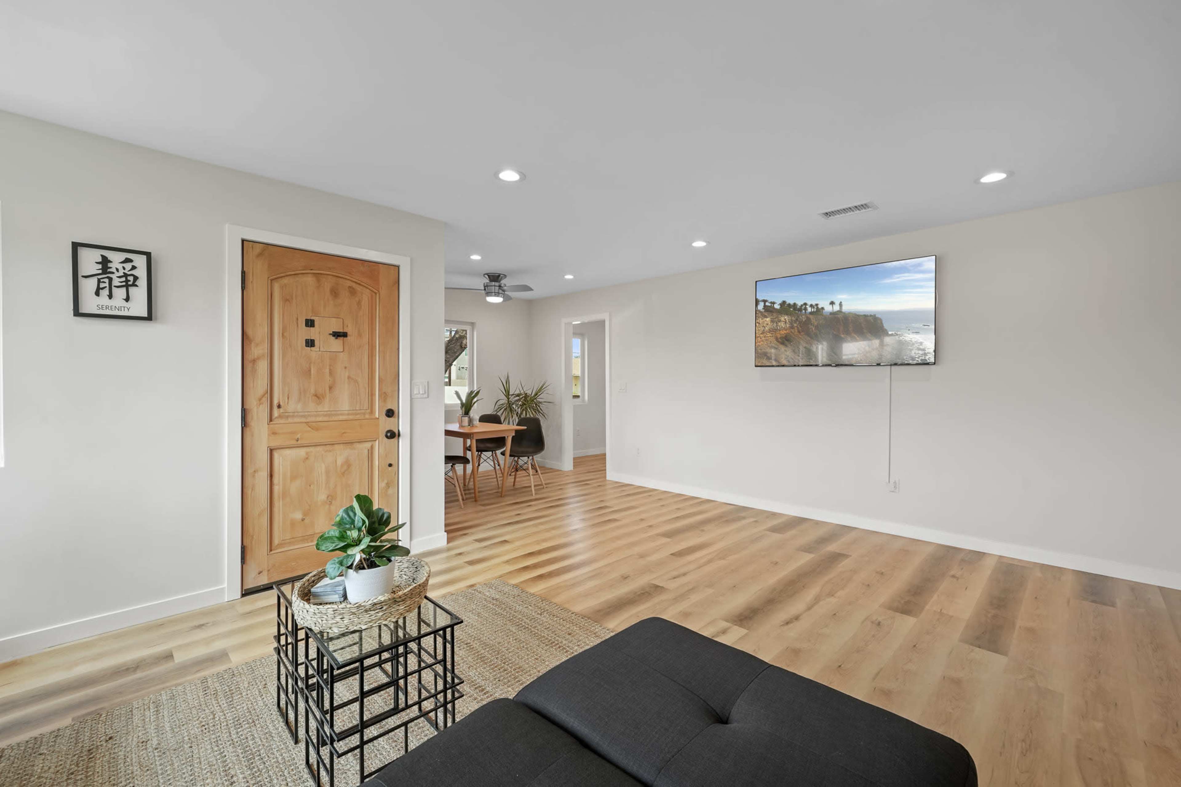 A modern living area features a door, a wall-mounted television displaying a coastal view, a dining table, and a decorative plant.