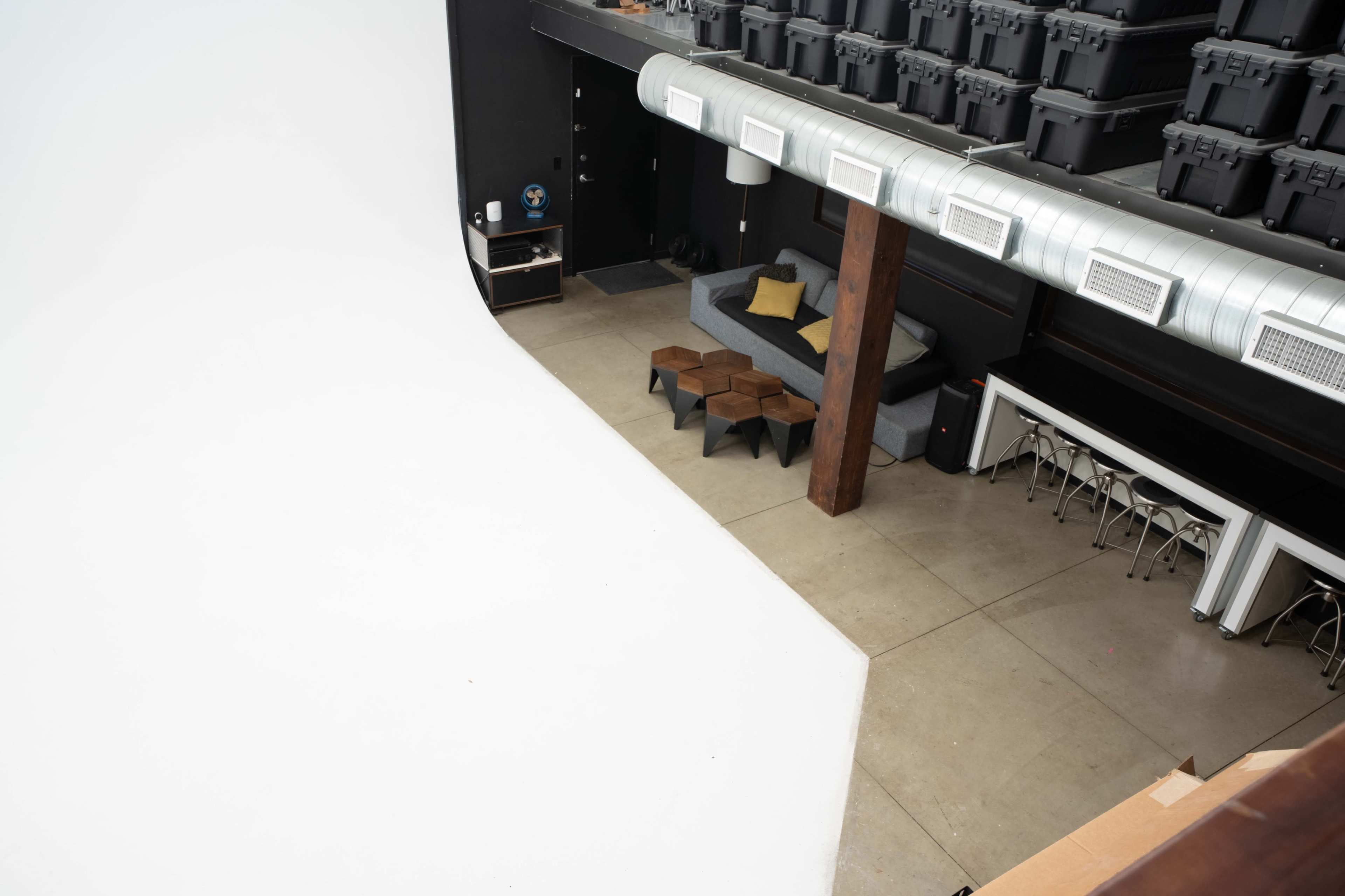 The image shows a spacious studio with a large white backdrop, a gray couch, stacked black storage boxes above, and modern furniture arranged on a concrete floor.