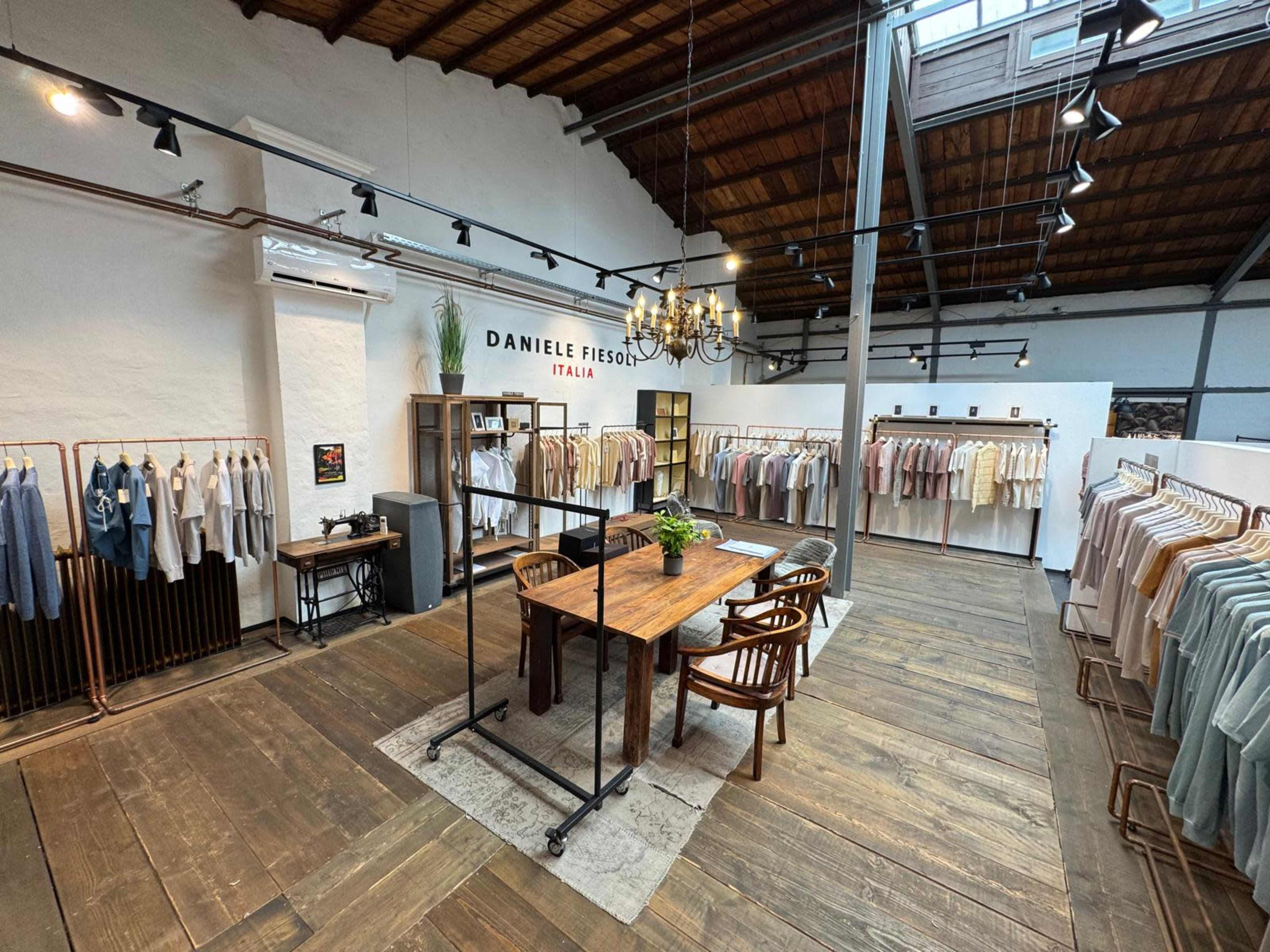 The image shows a clothing showroom with neatly arranged garments hanging on racks, a wooden table surrounded by chairs, and a warm, rustic interior design.