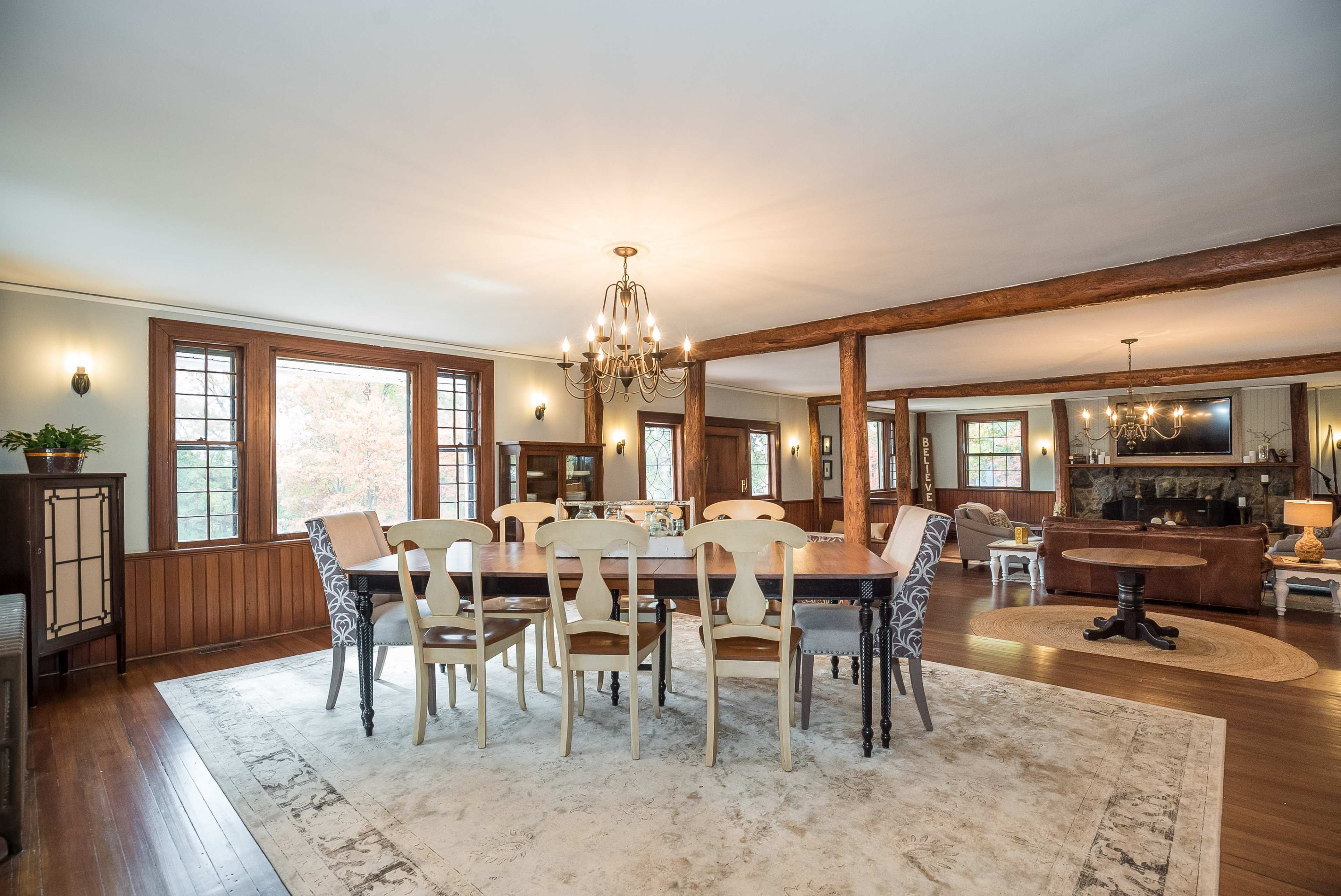 A spacious dining room featuring a long wooden table surrounded by mix-matched chairs, with large windows allowing natural light and a chandelier overhead.