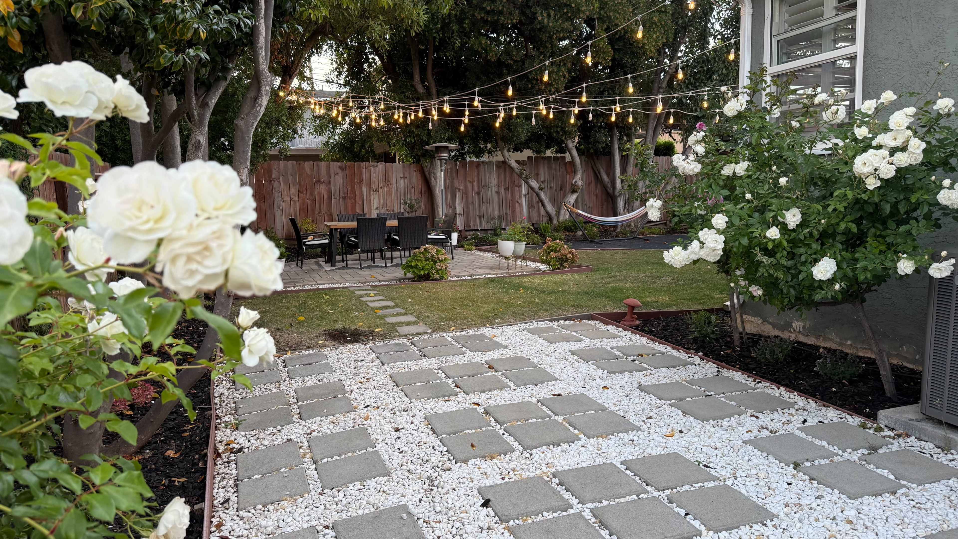 The image shows a landscaped backyard with white roses, a stone pathway, and string lights overhead, creating an inviting outdoor space with a seating area and a hammock.