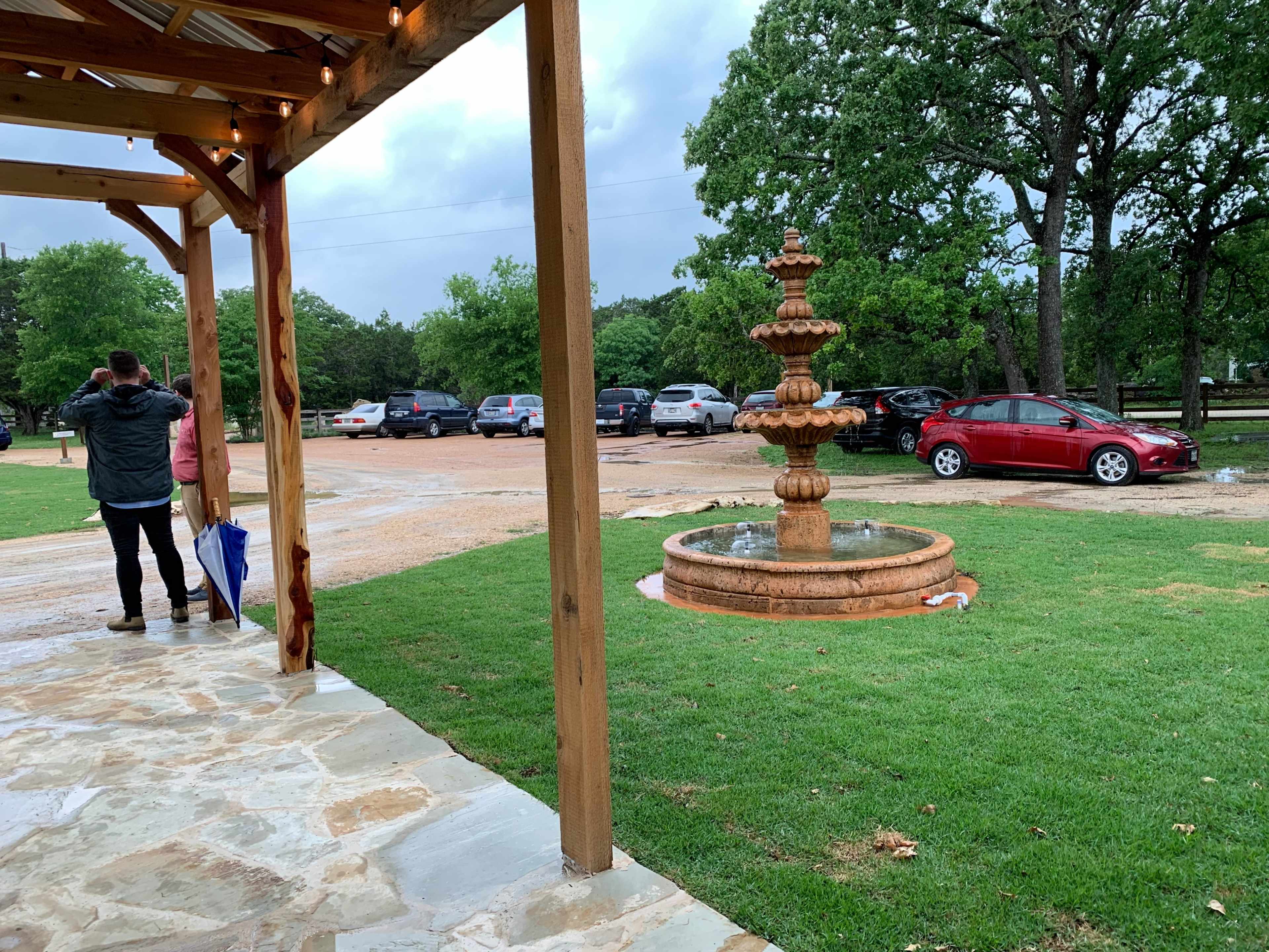 A fountain stands in the foreground as two people watch a parking area filled with cars under overcast skies.