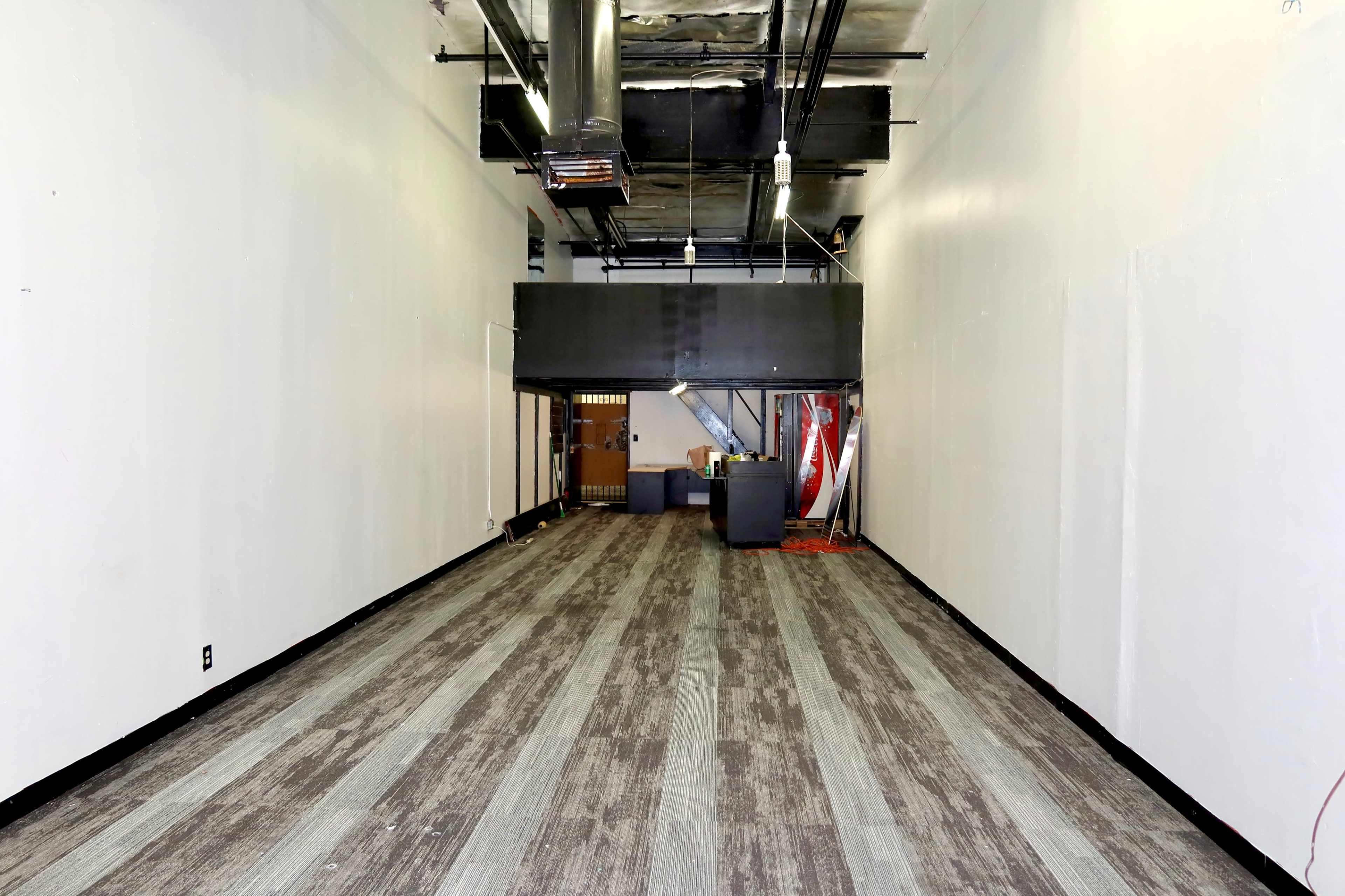 The image shows a long, empty hallway with a gray and brown striped carpet and white walls, leading to a staircase at the far end.