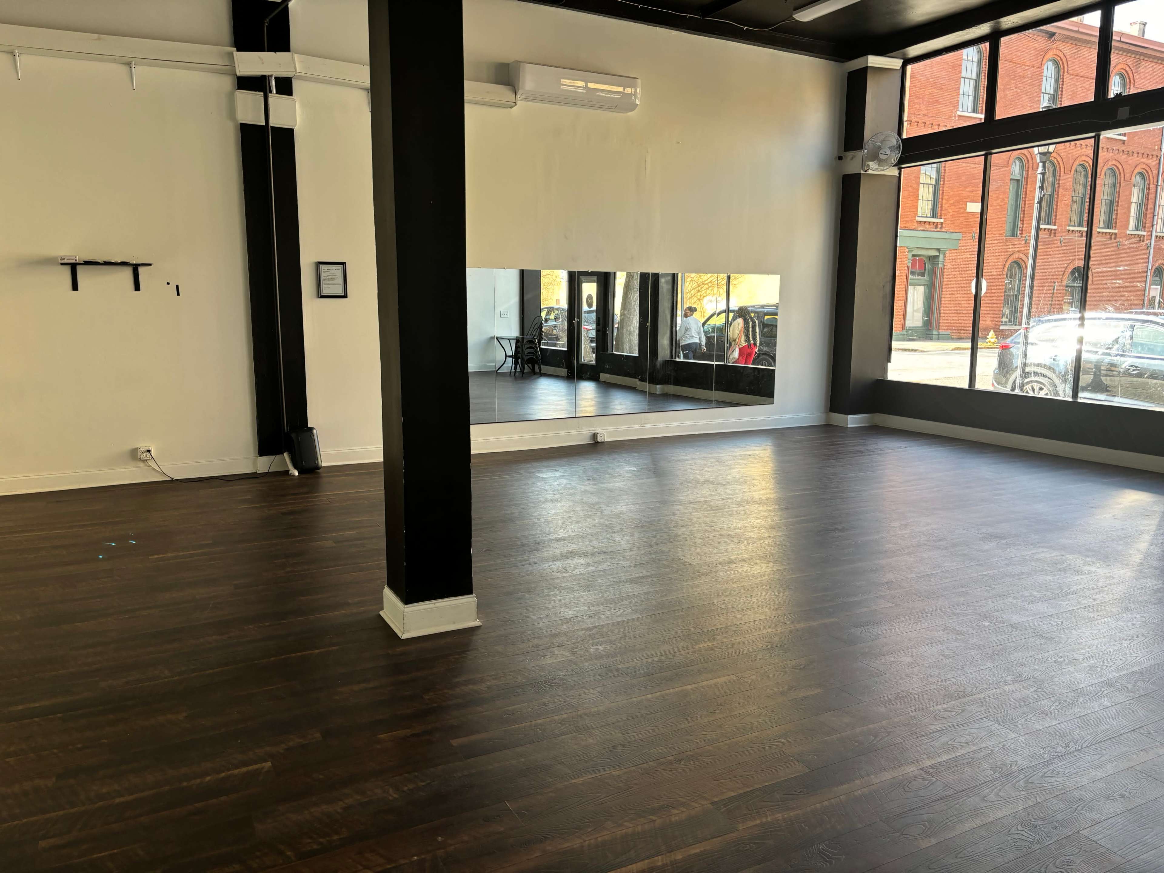 An empty interior space with dark wooden flooring, large windows, and a mirrored wall.
