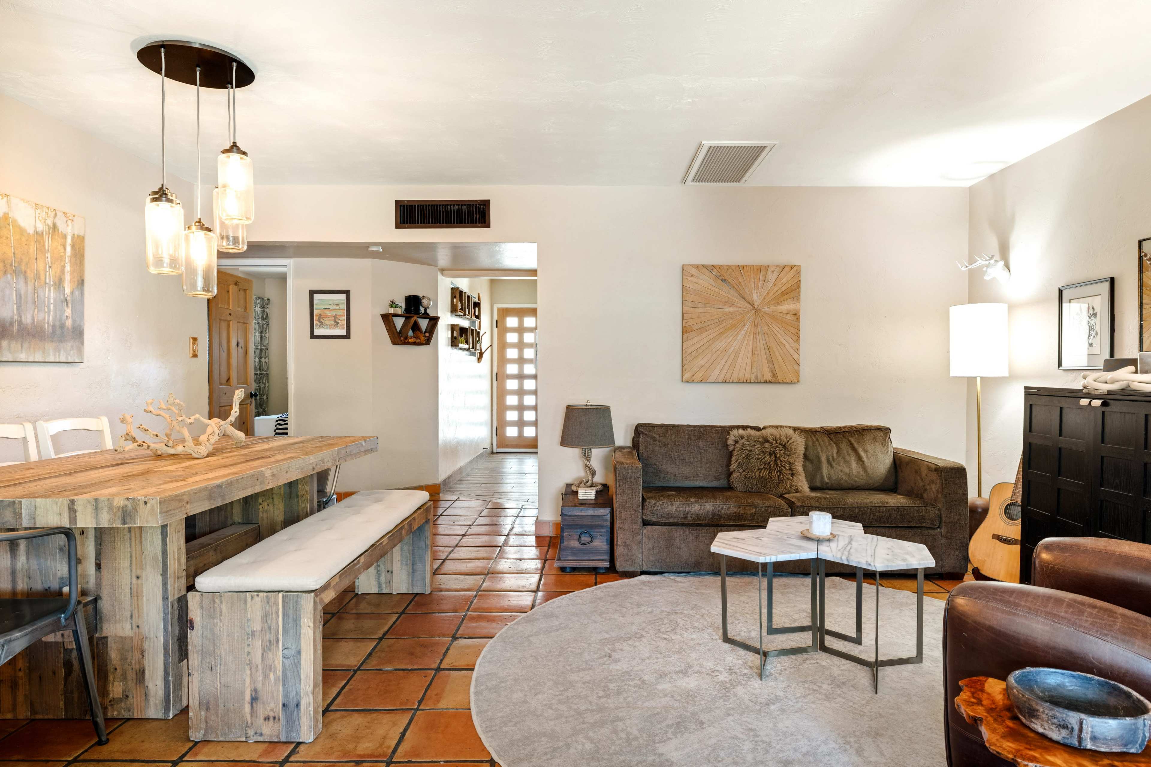 The image shows a cozy living room with a wooden dining table, a sofa, a coffee table, and a guitar resting against a chair, all set on a tiled floor.
