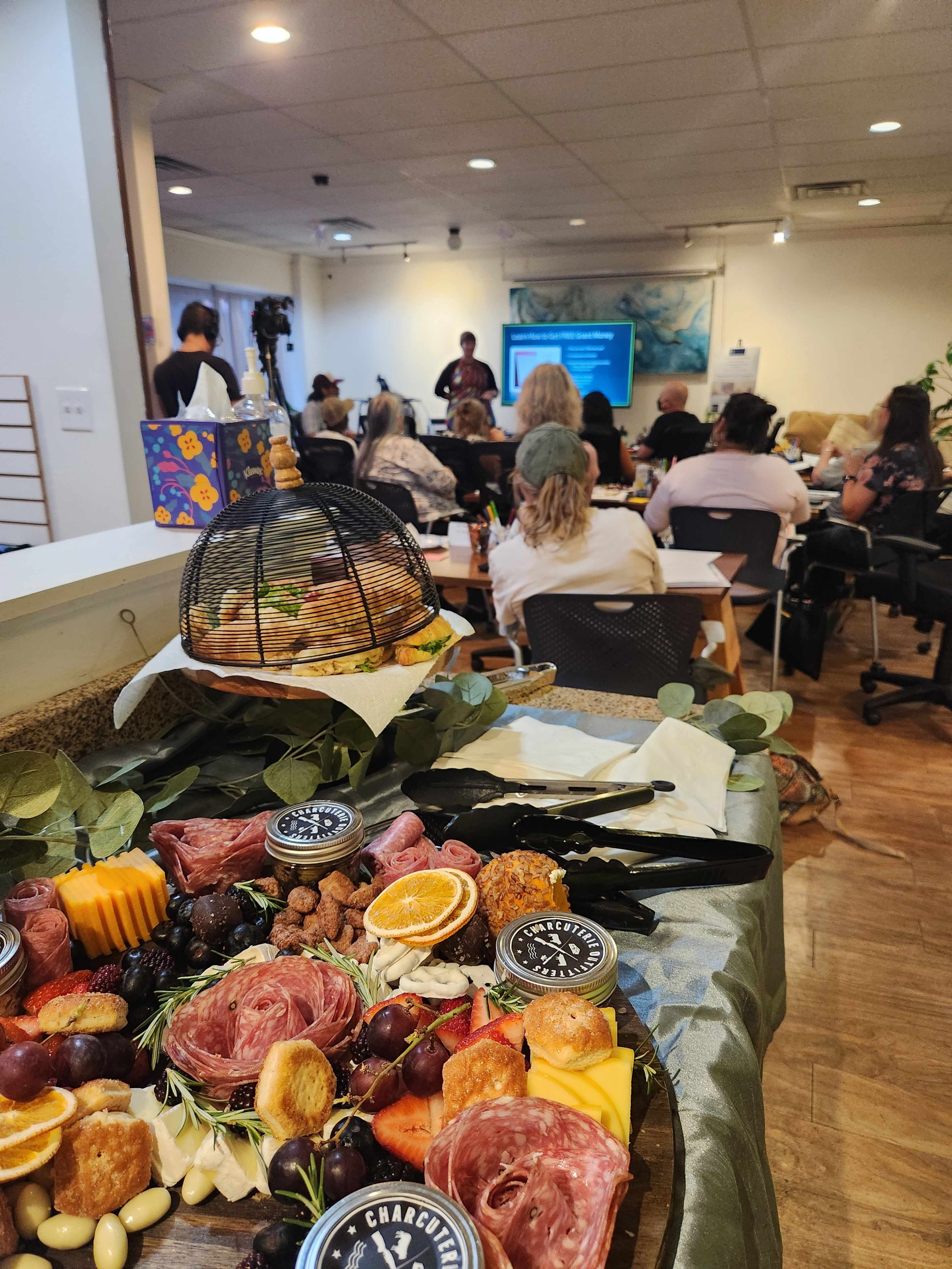 A variety of meats, cheeses, fruits, and nuts are arranged on a platter in the foreground, while a group of people listens to a presentation in a conference room in the background.
