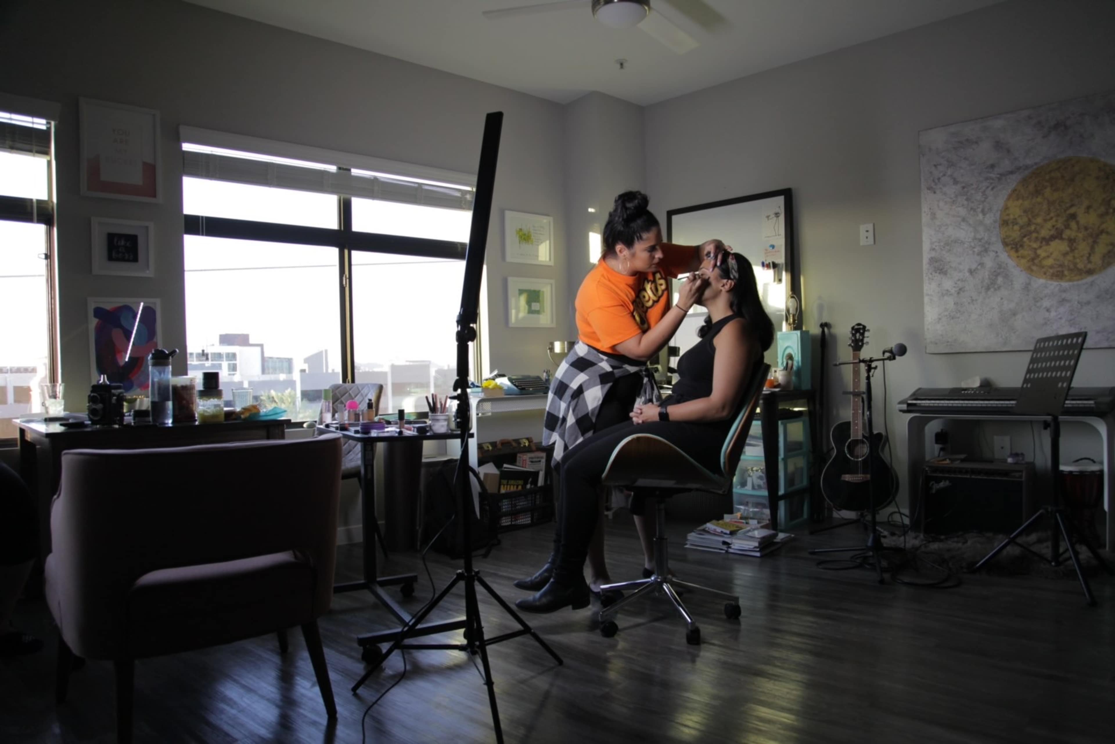 A makeup artist applies makeup to a client in a well-lit room with studio equipment and a desk filled with beauty products.