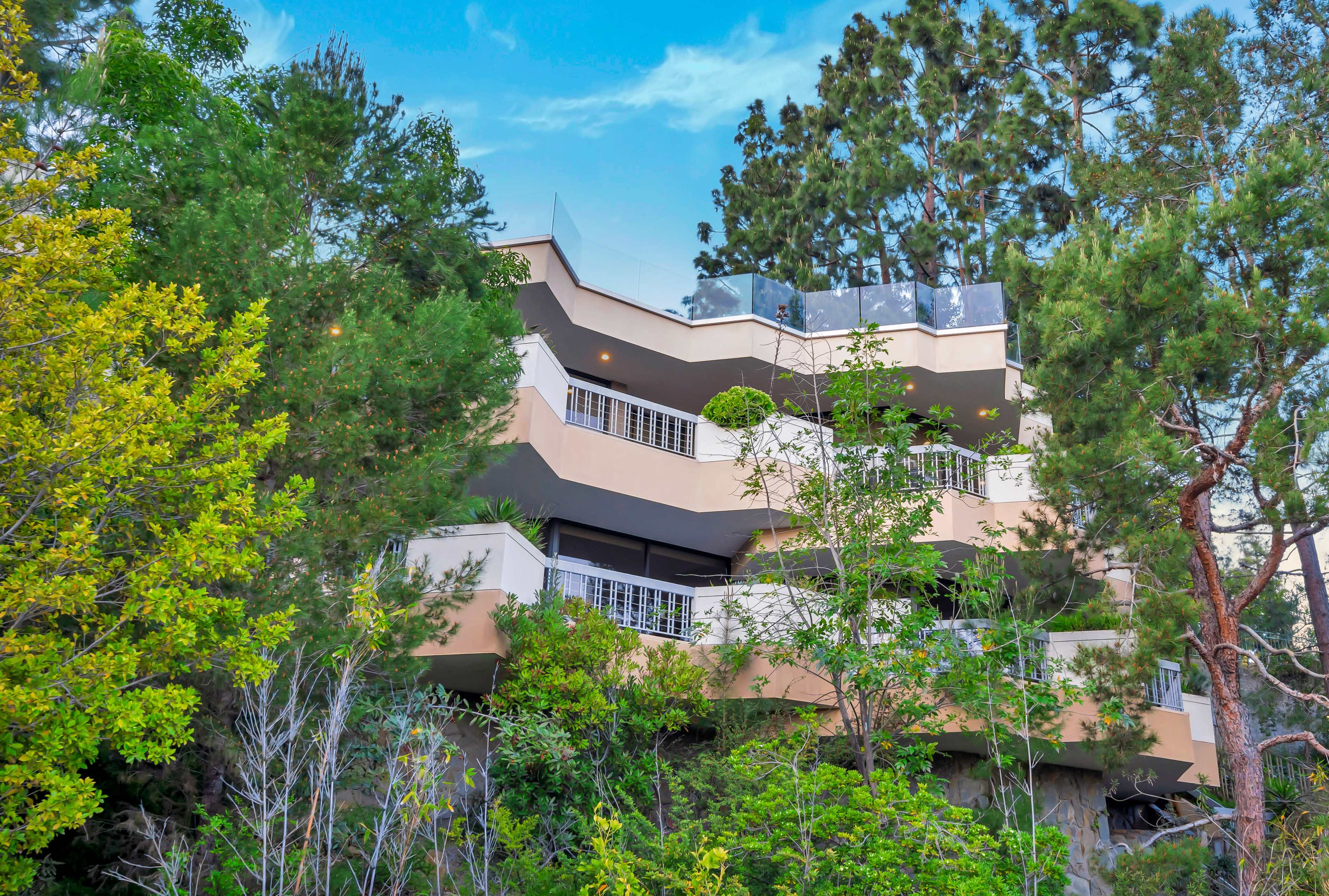 A multi-level modern house is nestled among lush green trees, featuring multiple balconies and a glass railing.