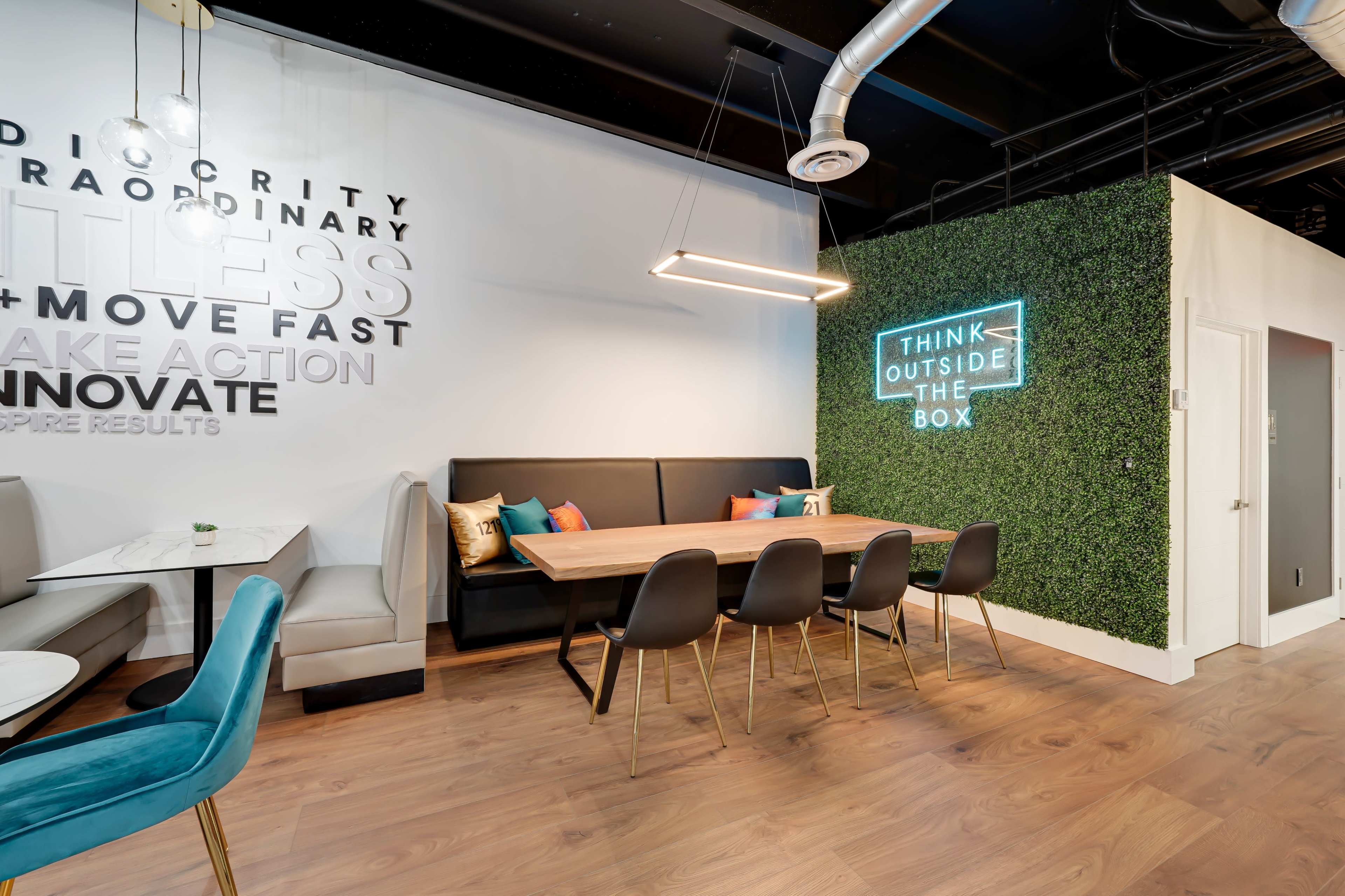 A modern workspace features a large wooden table surrounded by black chairs, with wall decor that includes inspirational phrases and a green plant backdrop.