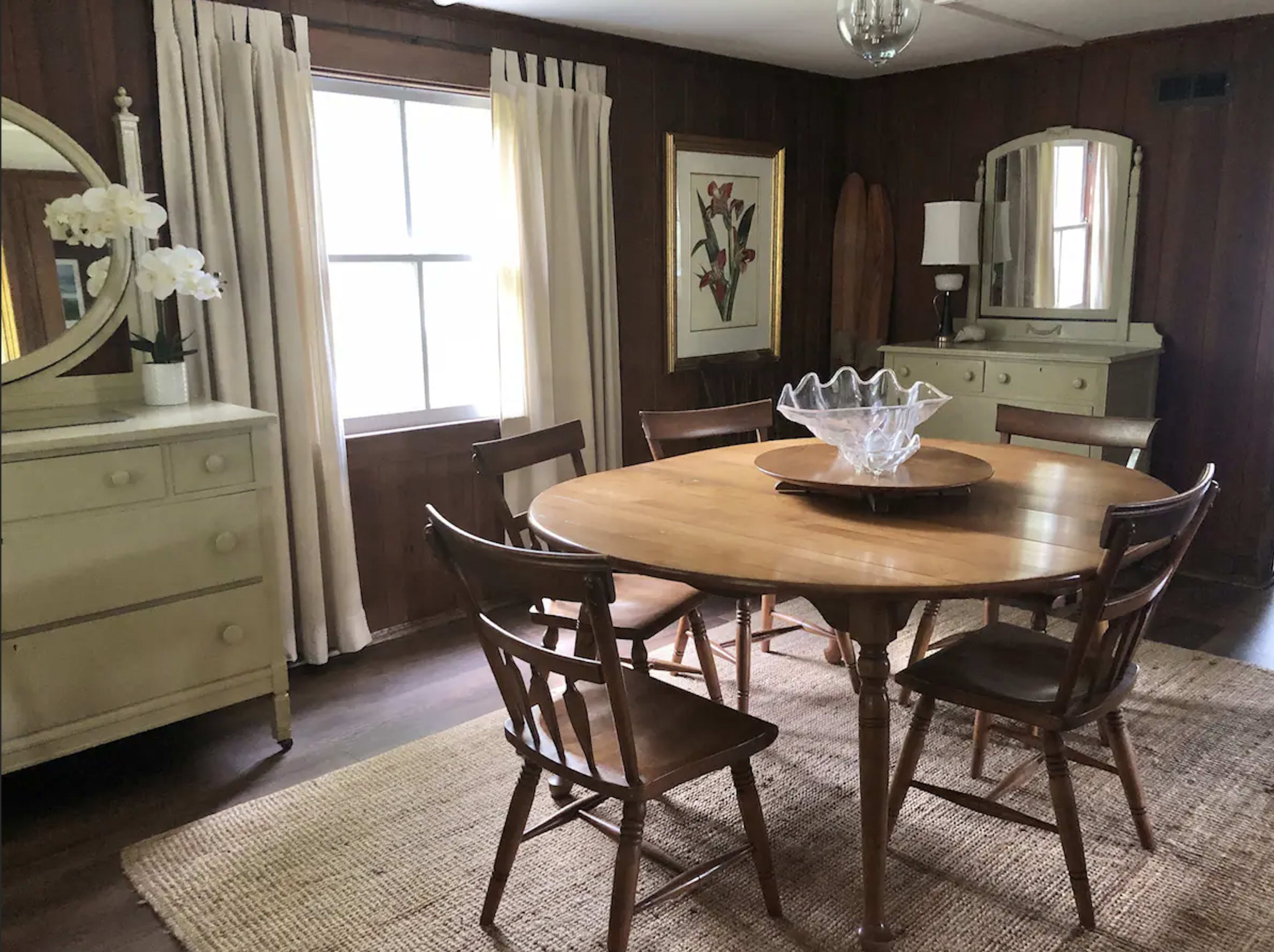 A round wooden dining table surrounded by four chairs sits in a room with wooden walls, large windows, and light-colored furniture.