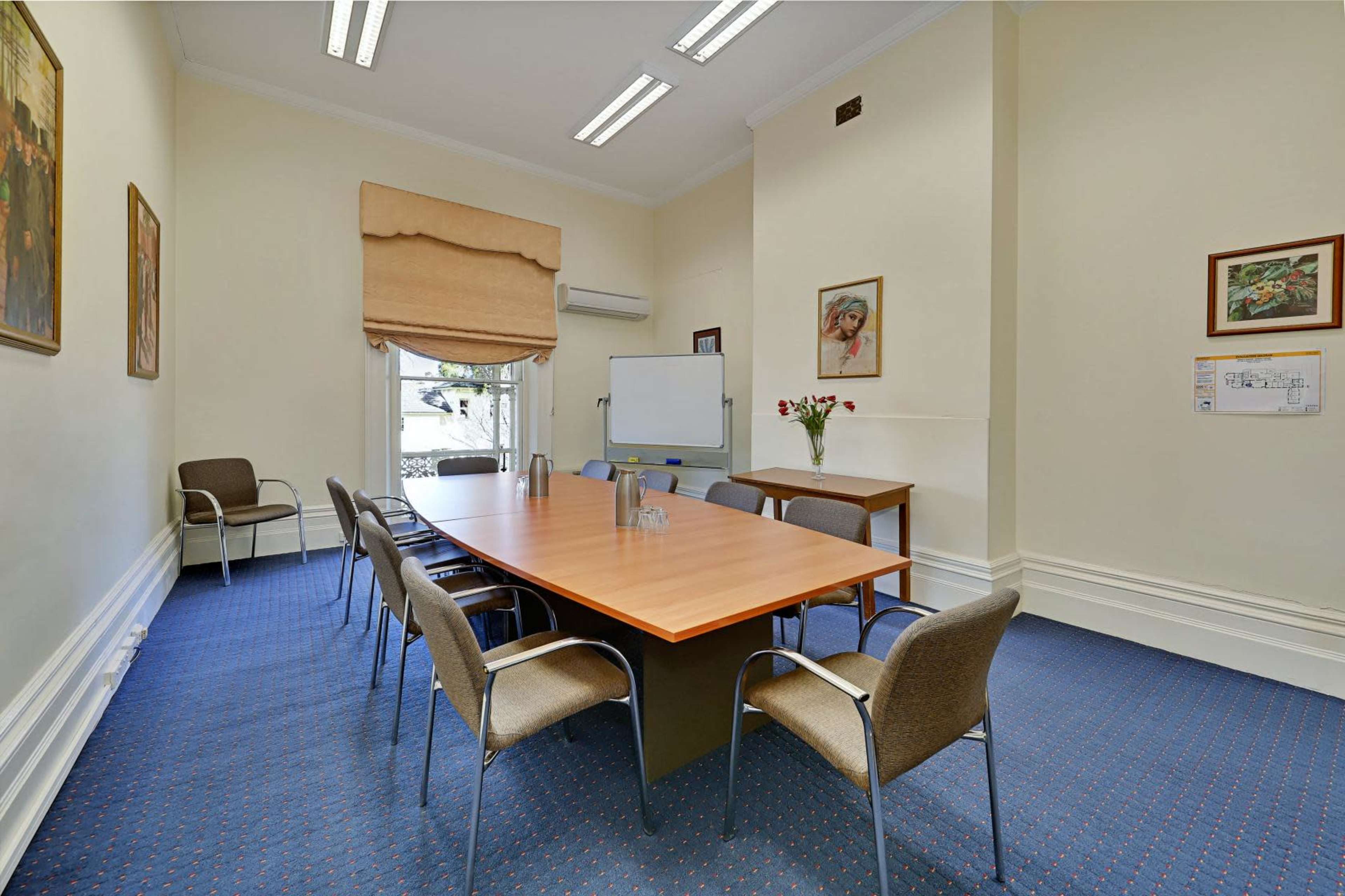 The image shows a meeting room with a wooden table surrounded by eight chairs, a whiteboard, and a window with a view.