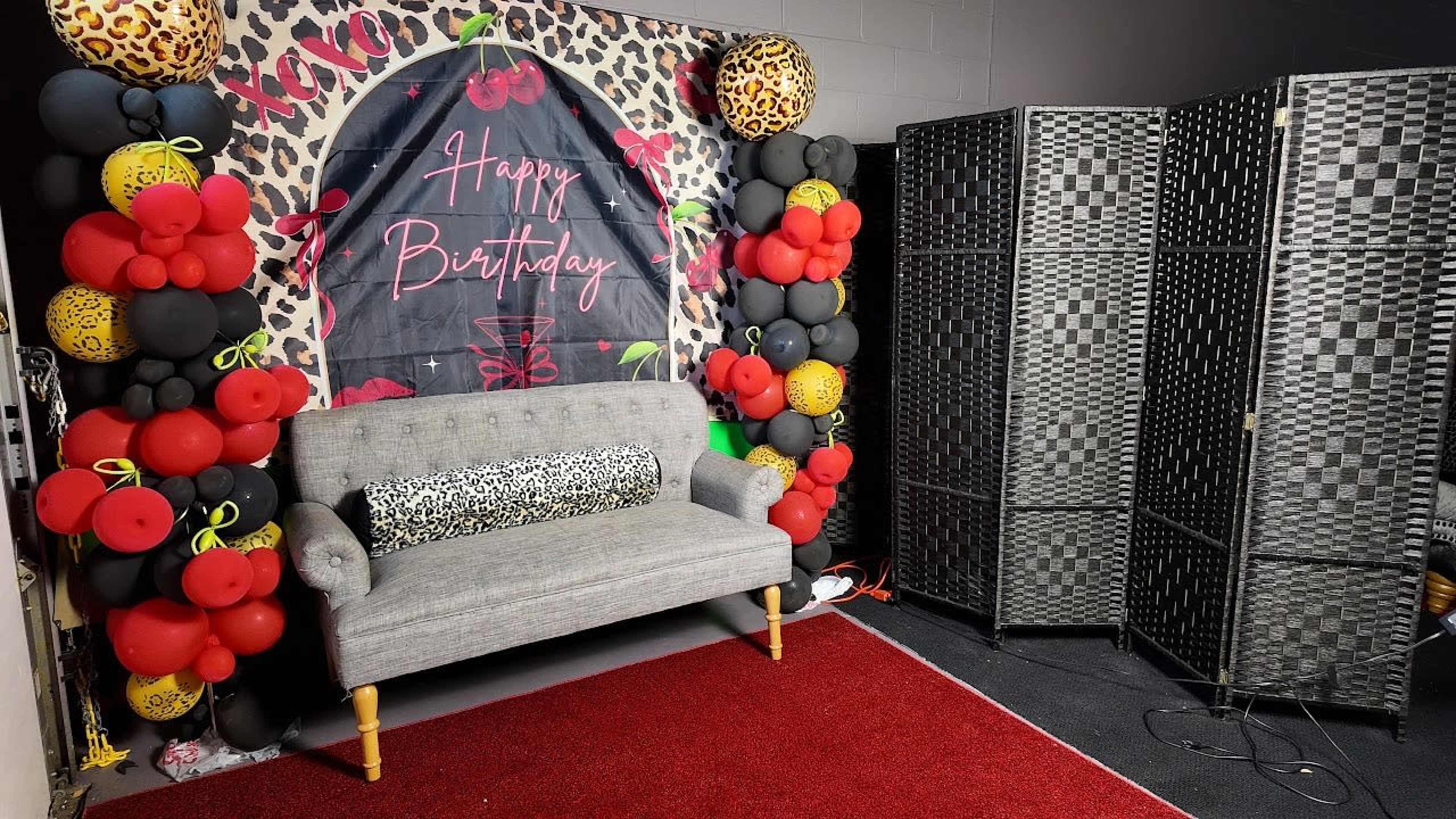 The image shows a decorated birthday photo backdrop featuring a black and leopard print banner with balloons and a grey couch in front, alongside a folding screen.