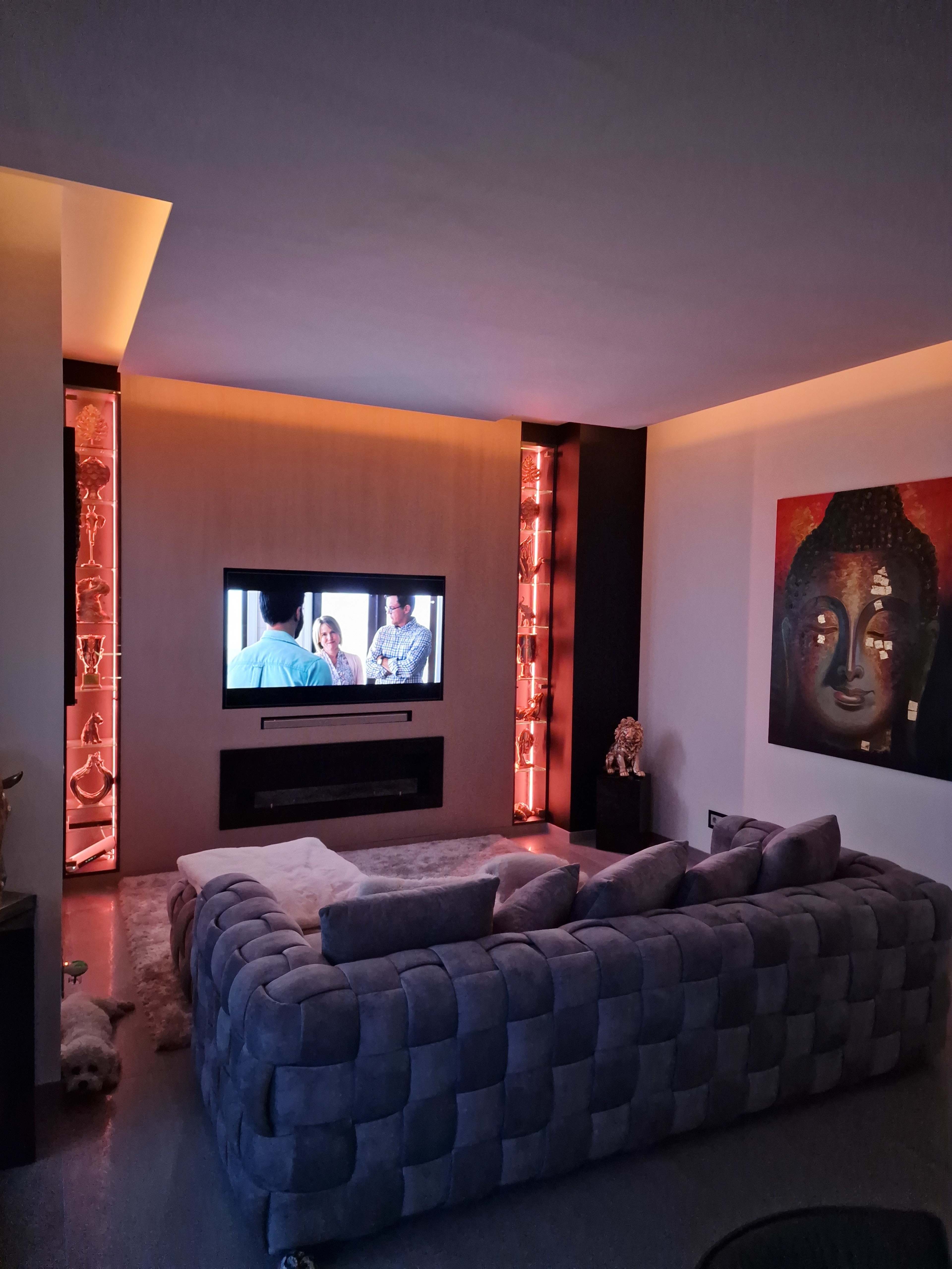 A modern living room with a large gray sofa facing a wall-mounted TV, surrounded by decorative shelves with ambient lighting and a large Buddha painting on the adjacent wall.