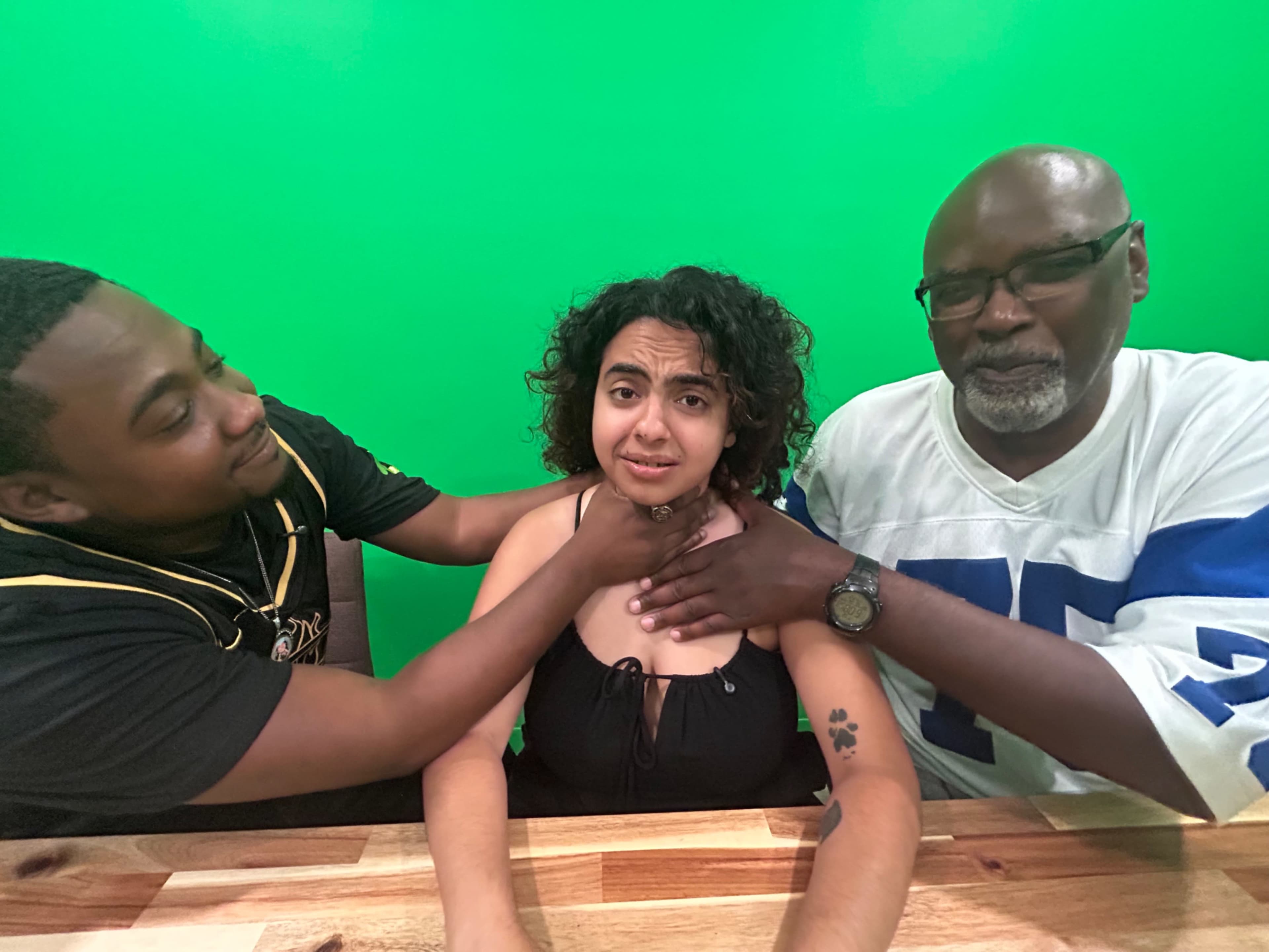 Two men are playfully interacting with a woman at a wooden table in front of a green screen backdrop.