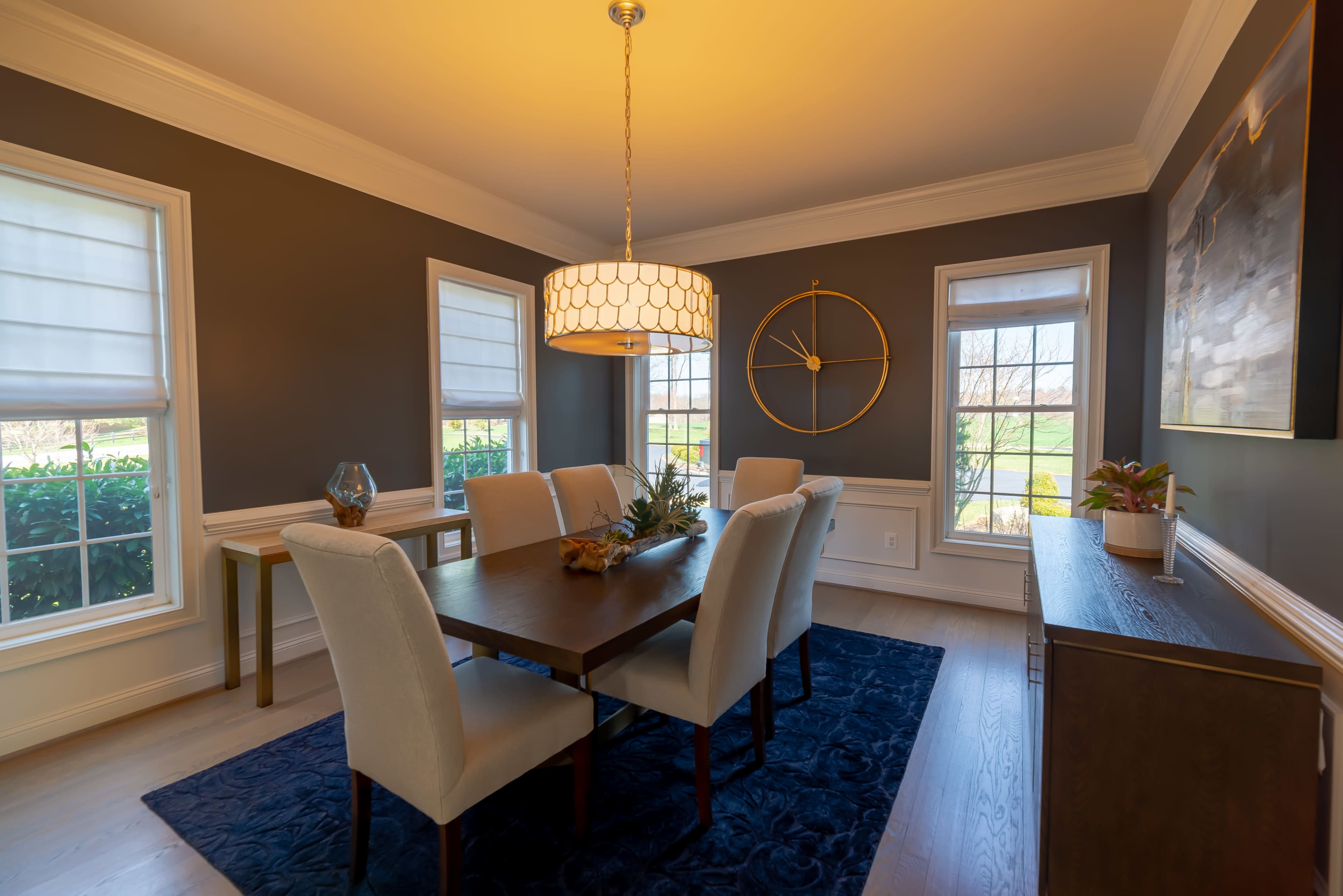 A dining room features a wooden table surrounded by six upholstered chairs, with a pendant light overhead and windows allowing natural light to enter.