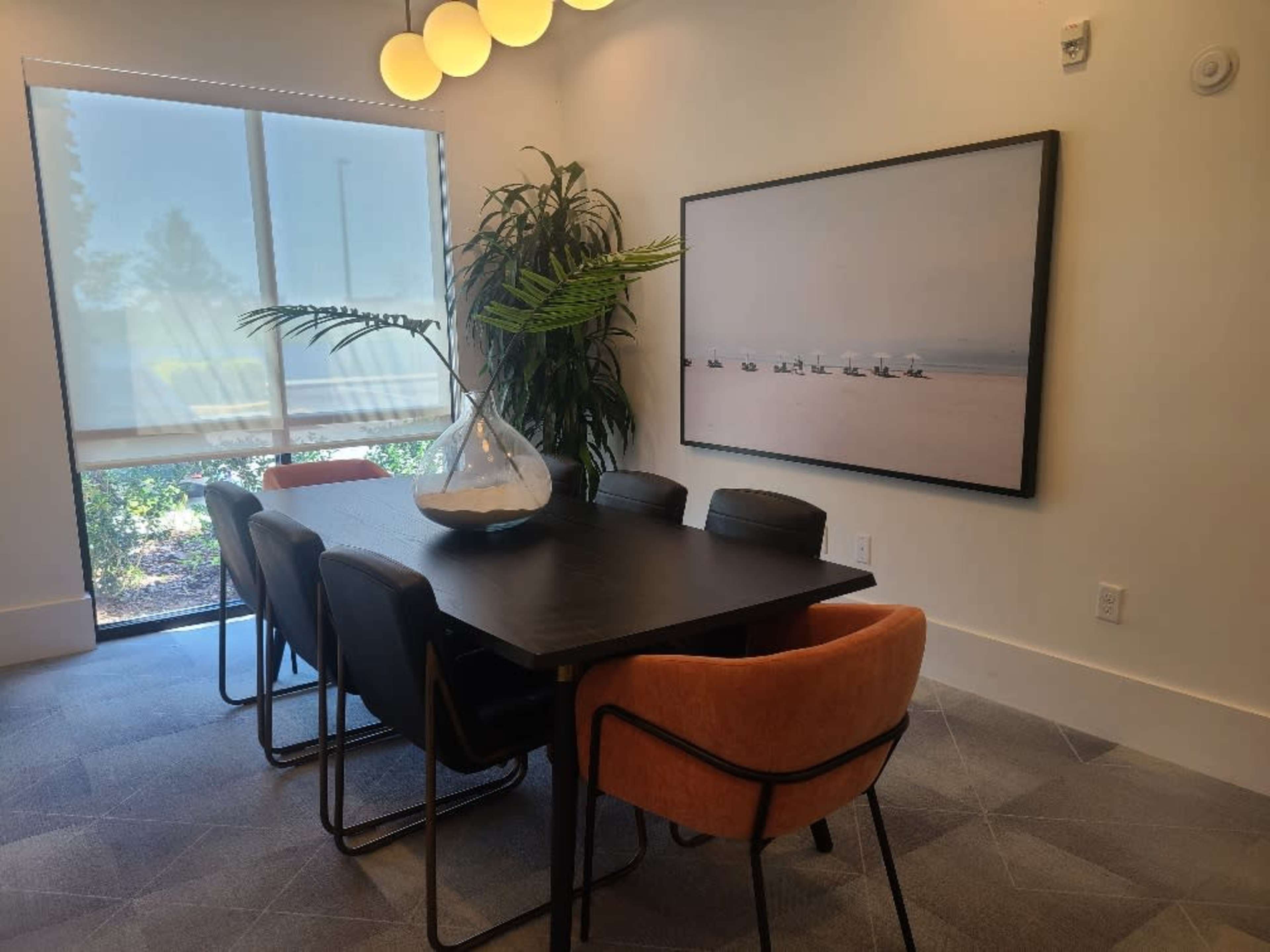 A modern conference room features a large table surrounded by black and orange chairs, with a plant and a decorative vase on the table, and a large framed artwork on the wall.