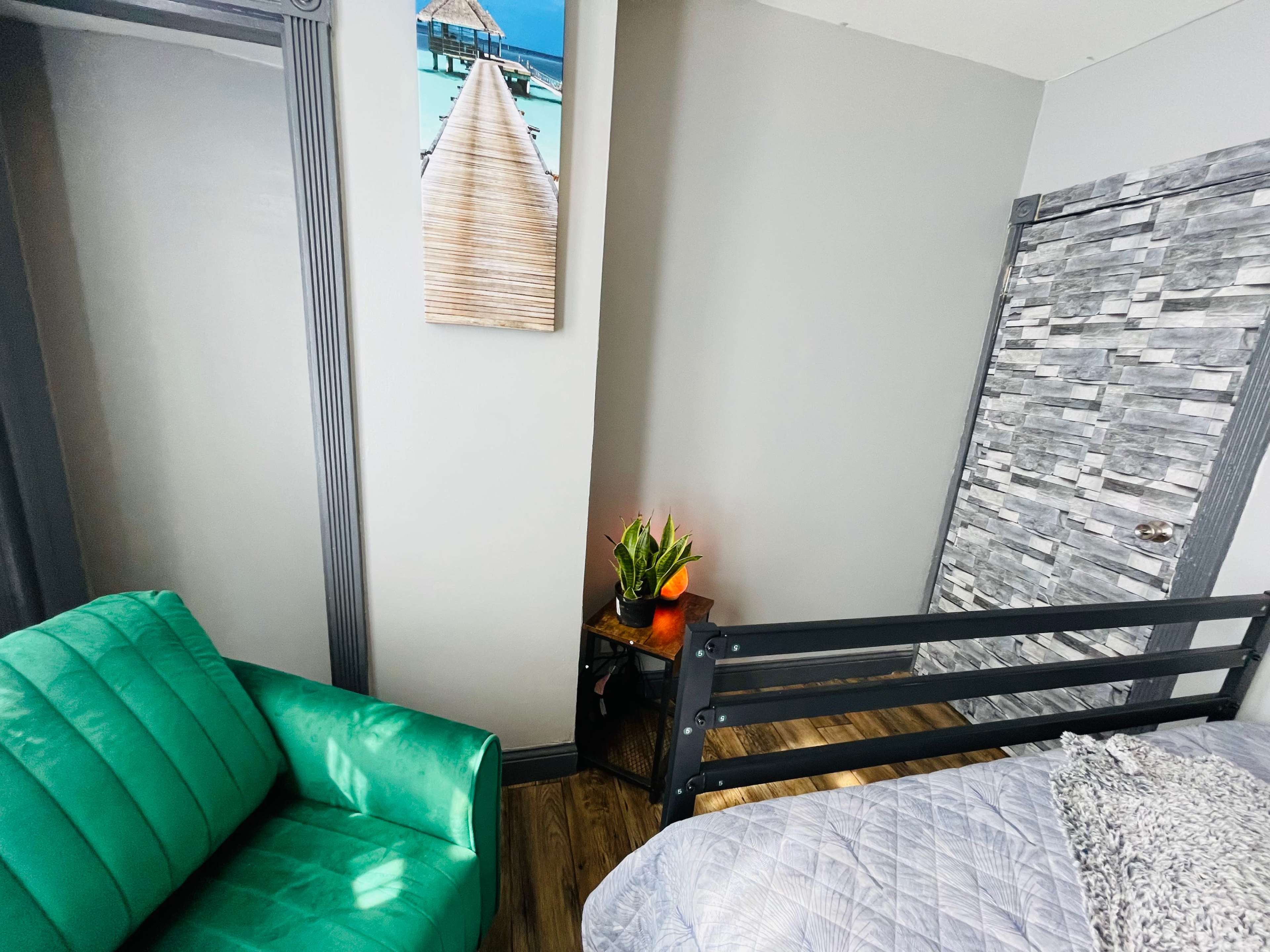 A corner of a room featuring a green couch, a bedside with a potted plant, and a bed with gray bedding against a wall with a decorative wooden panel.