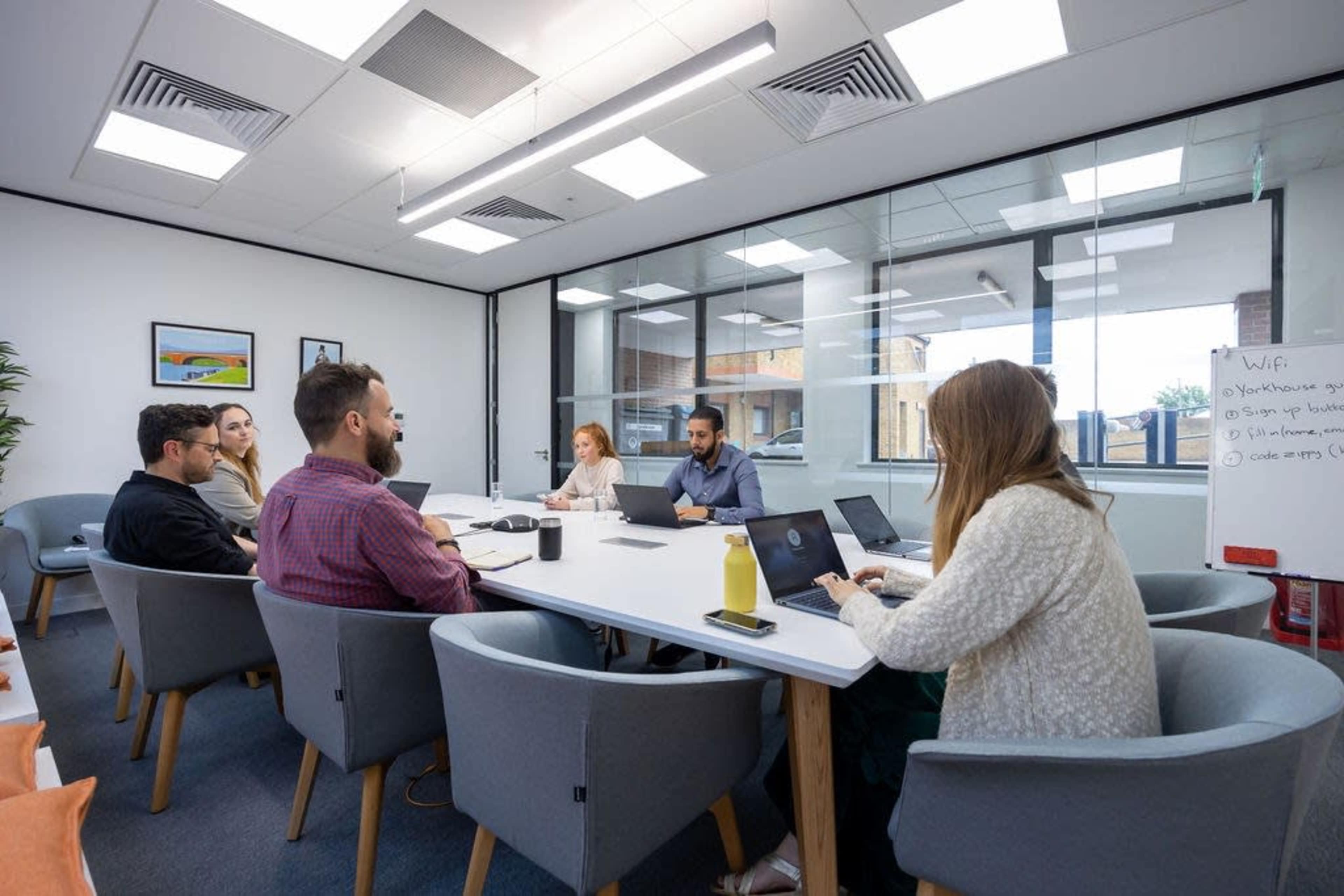 A group of six people sit around a large conference table in a well-lit meeting room, working on laptops and discussing ideas.