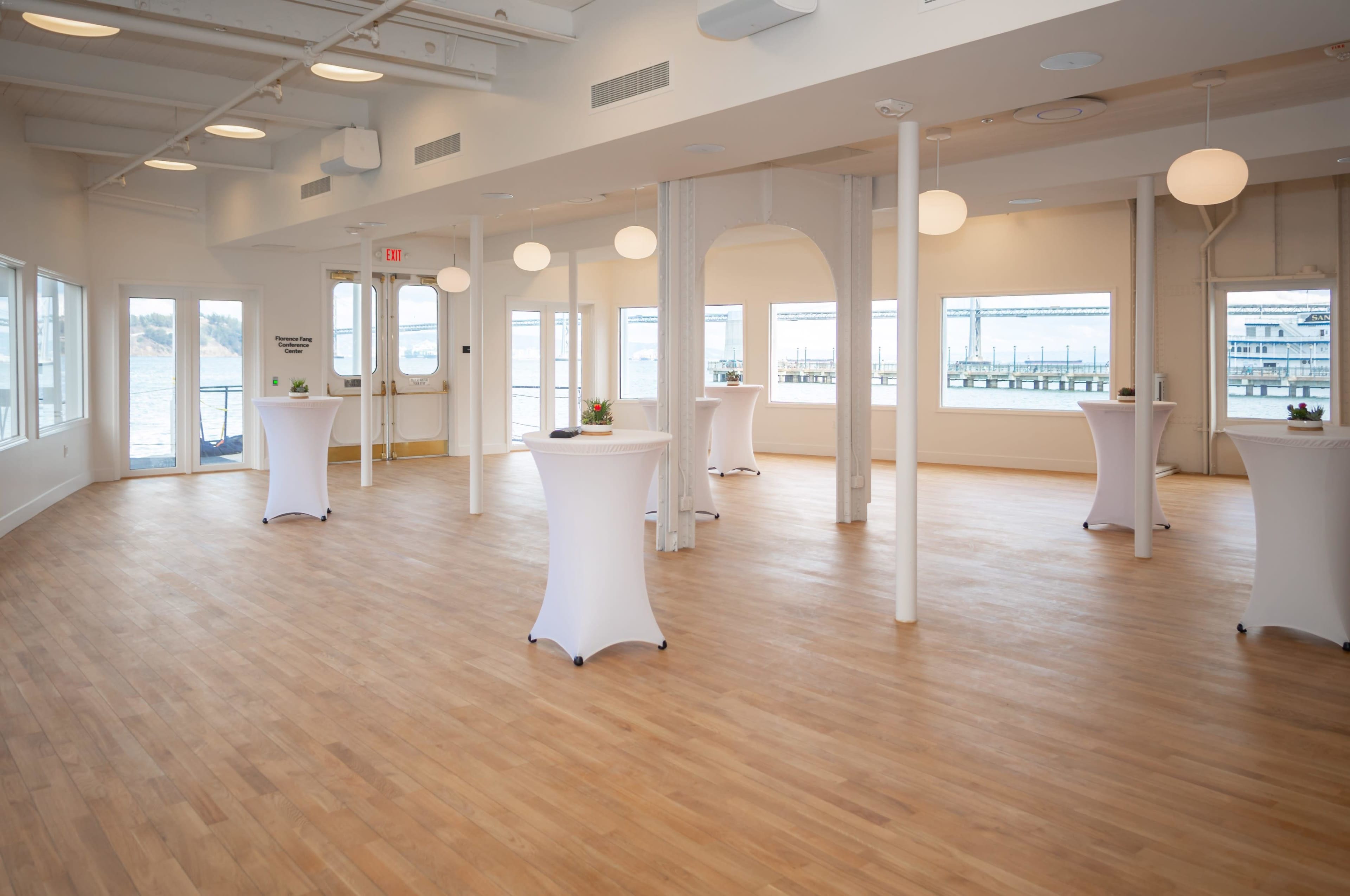 A spacious, well-lit event room with wooden flooring and several cocktail tables arranged around the space.