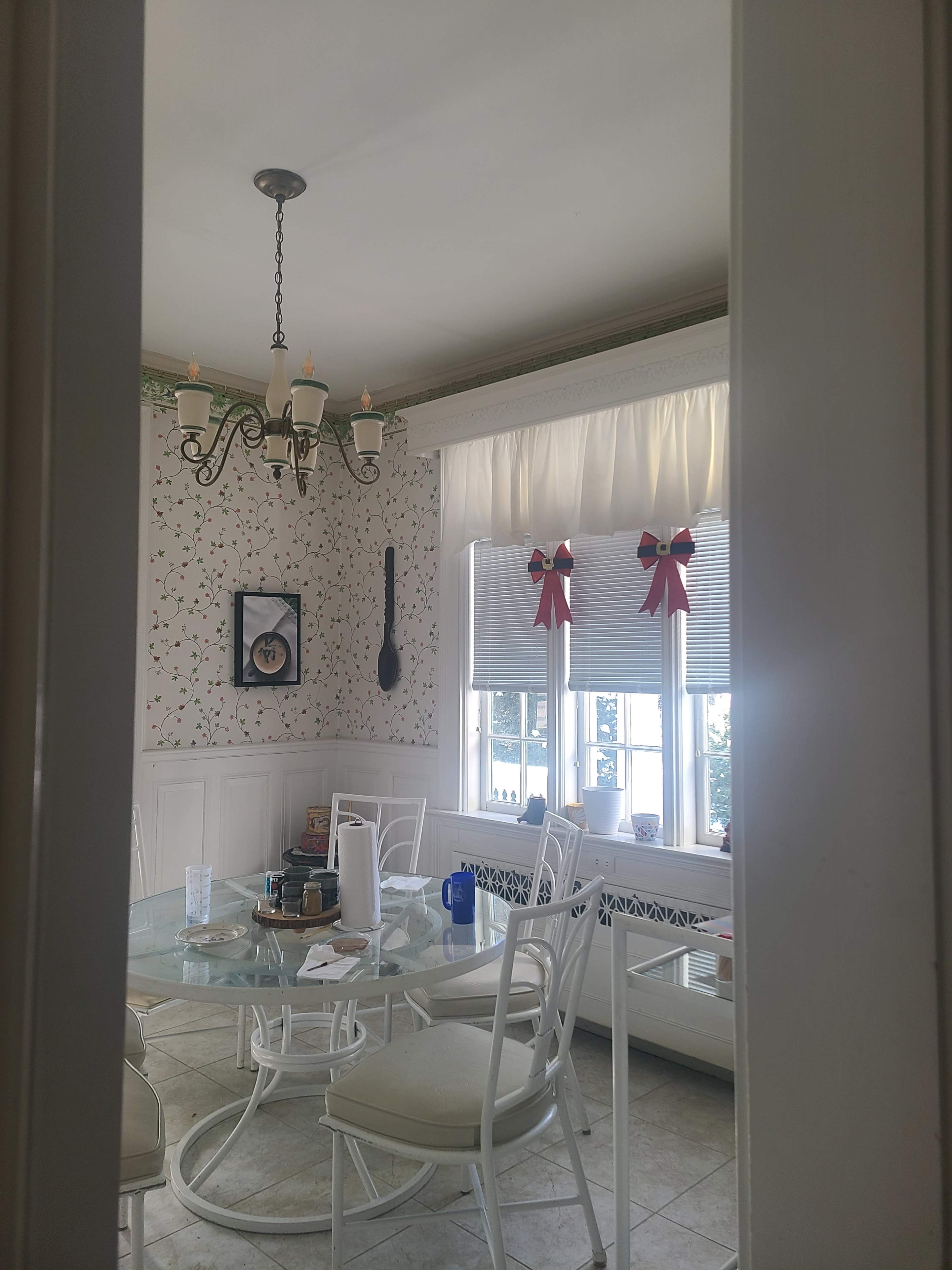 The image shows a dining room with a glass table and white chairs, decorated with a floral wallpaper and curtains adorned with red bows.