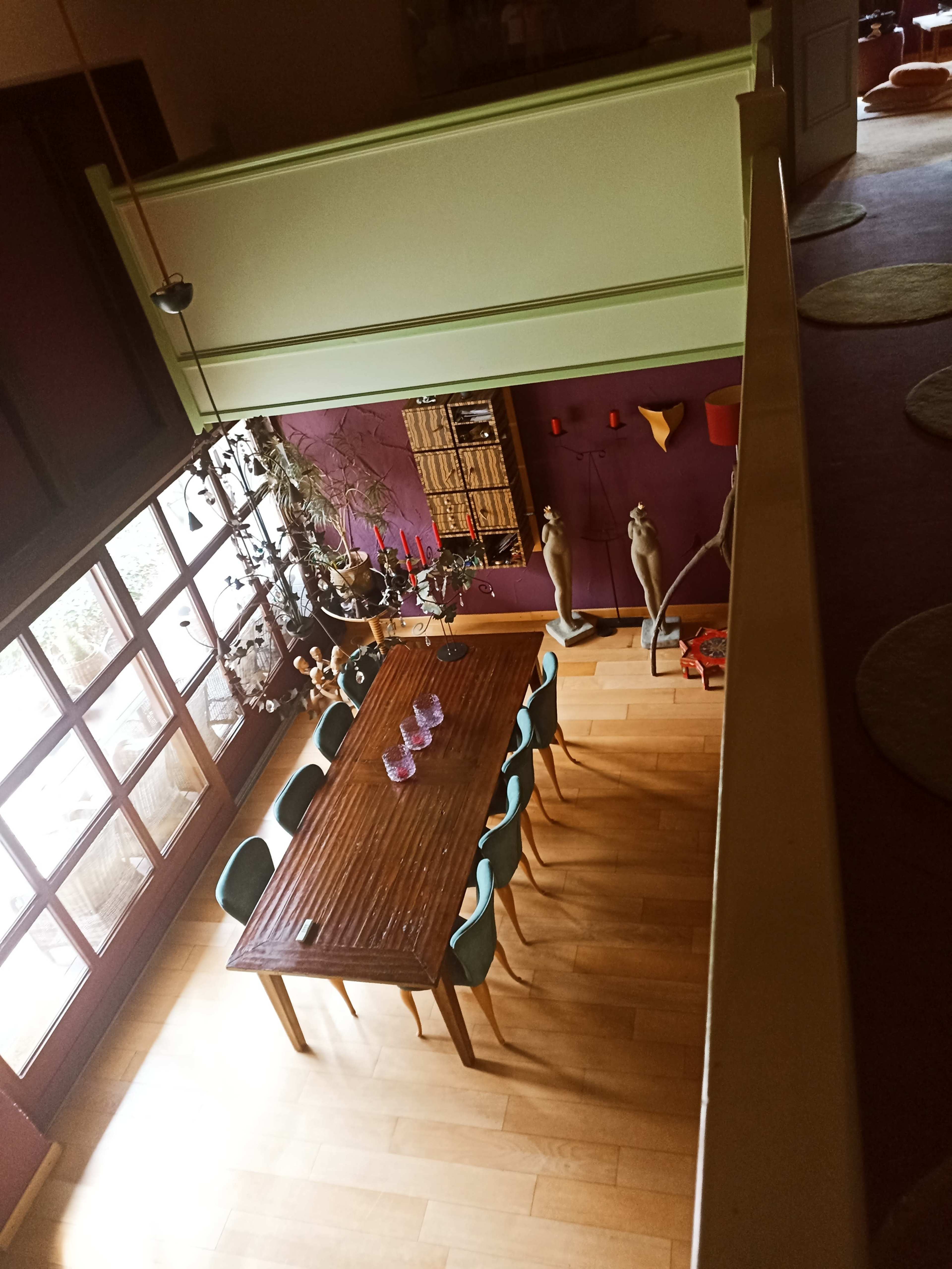 The image shows a spacious dining area with a wooden table surrounded by green chairs, illuminated by natural light from large windows.