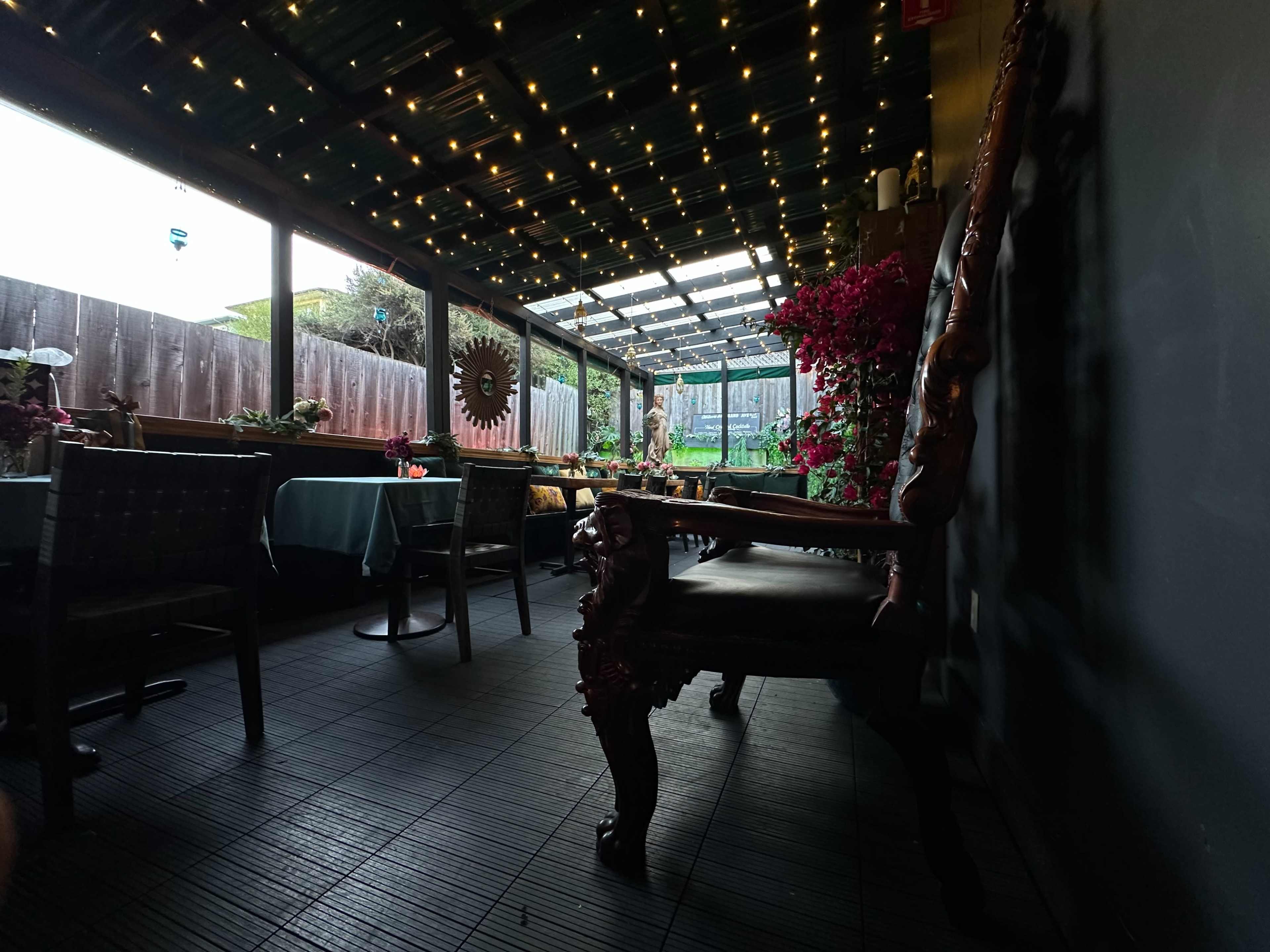 The image shows a dining area with decorated tables and a partially covered ceiling adorned with string lights, surrounded by plants and a wooden fence.