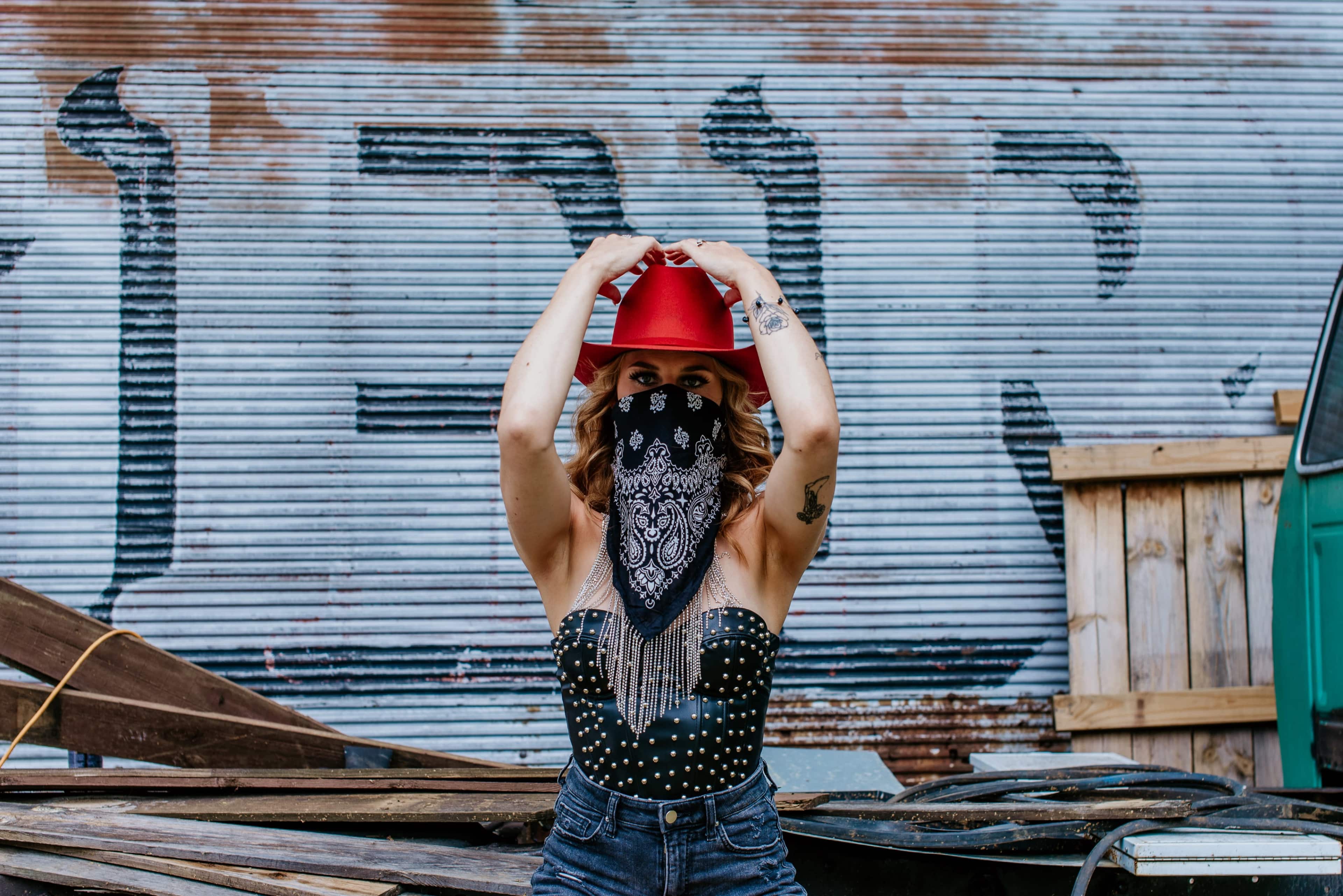 A person wearing a red hat and a black bandana stands in front of a corrugated metal background with graffiti.