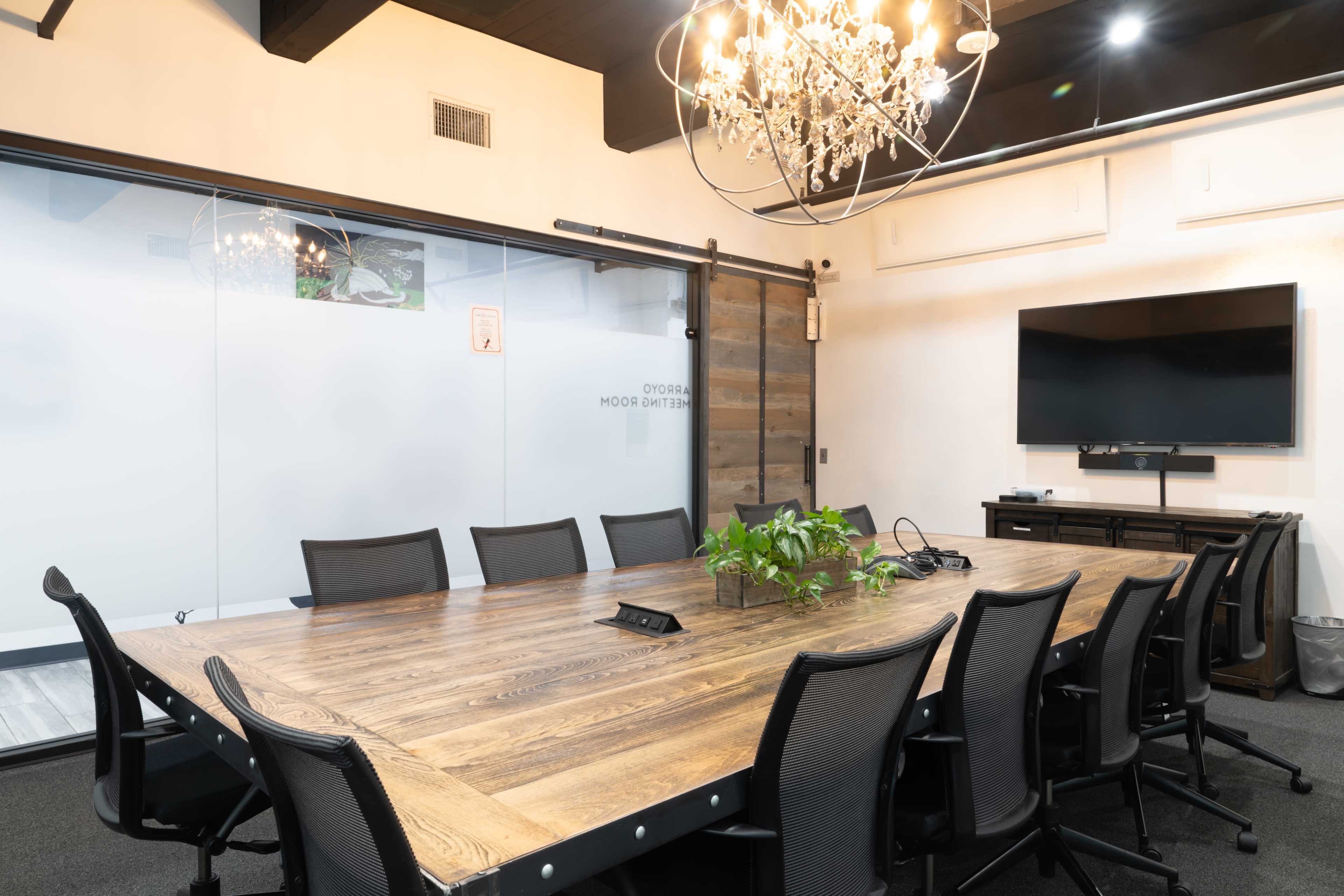 The image shows a modern conference room featuring a long wooden table surrounded by black task chairs, a large television mounted on the wall, and a chandelier overhead.