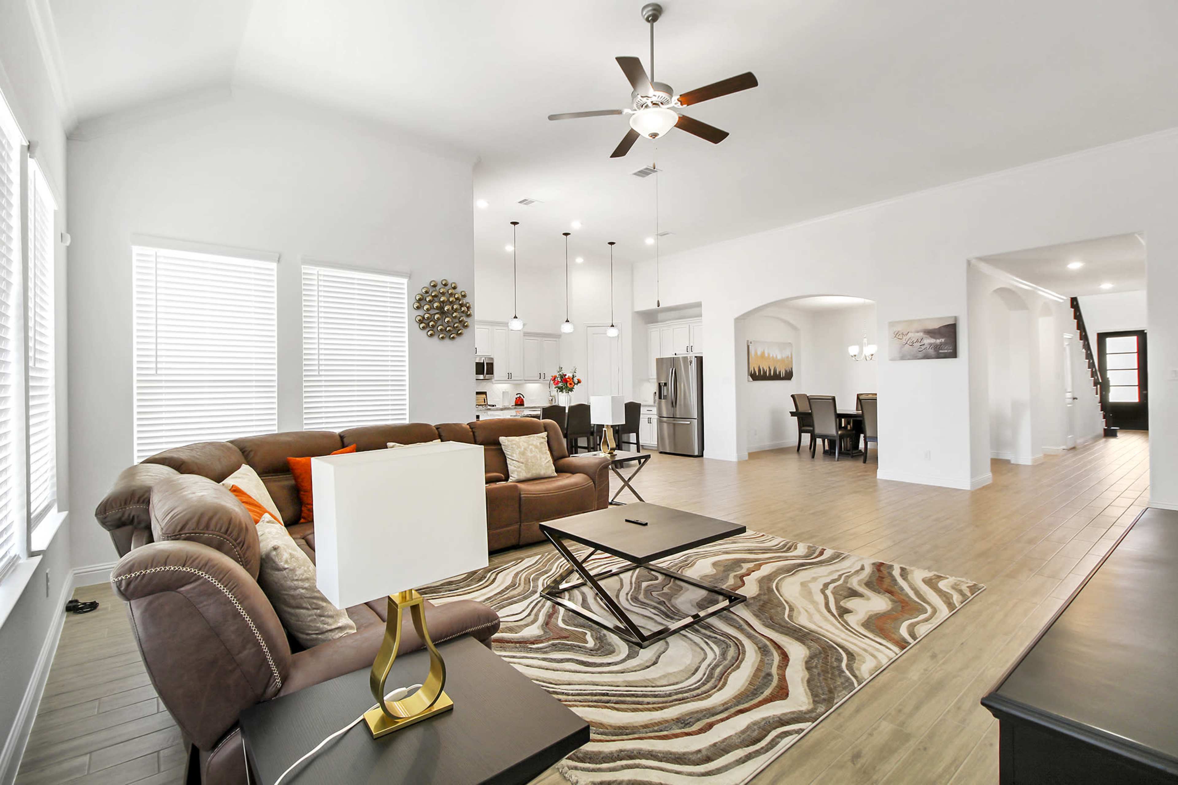 A spacious living room with a brown sectional sofa, a coffee table, and a large window allowing natural light, leading to an open kitchen and dining area.