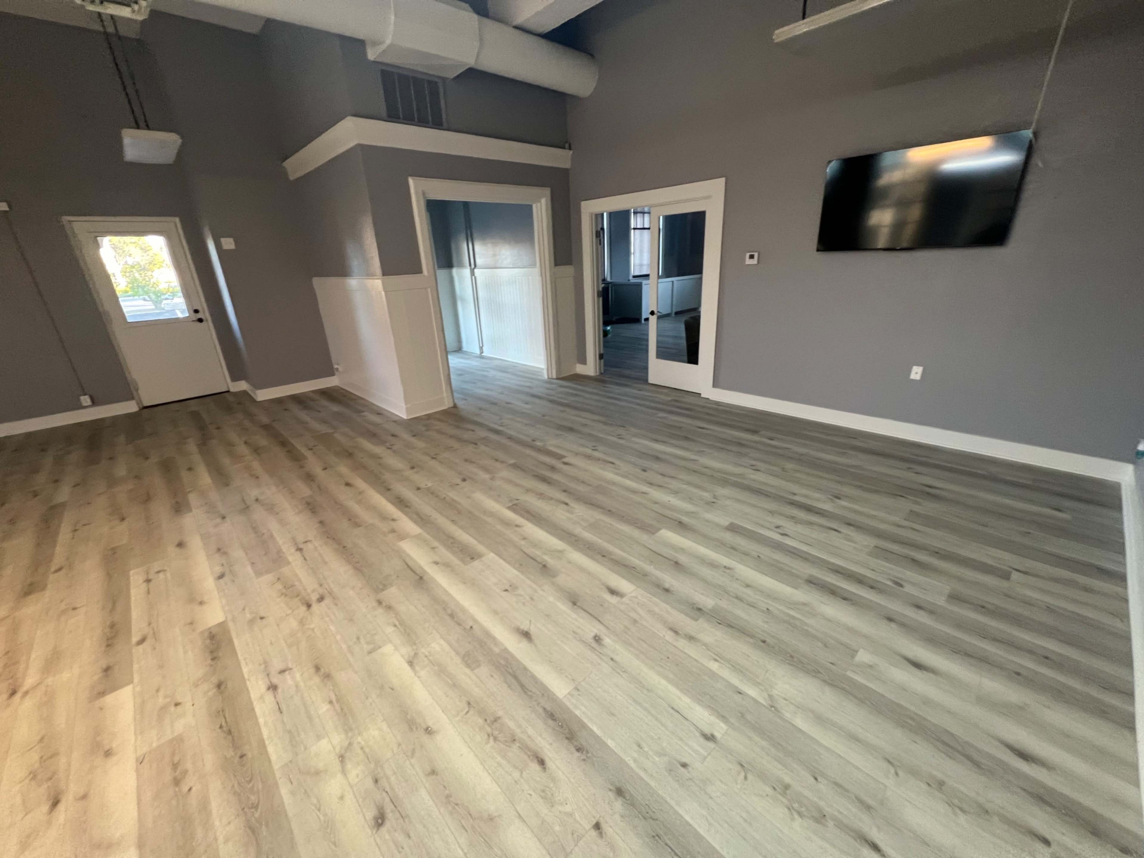 The image shows a spacious room with light hardwood flooring, gray walls, and a large television mounted on one wall.