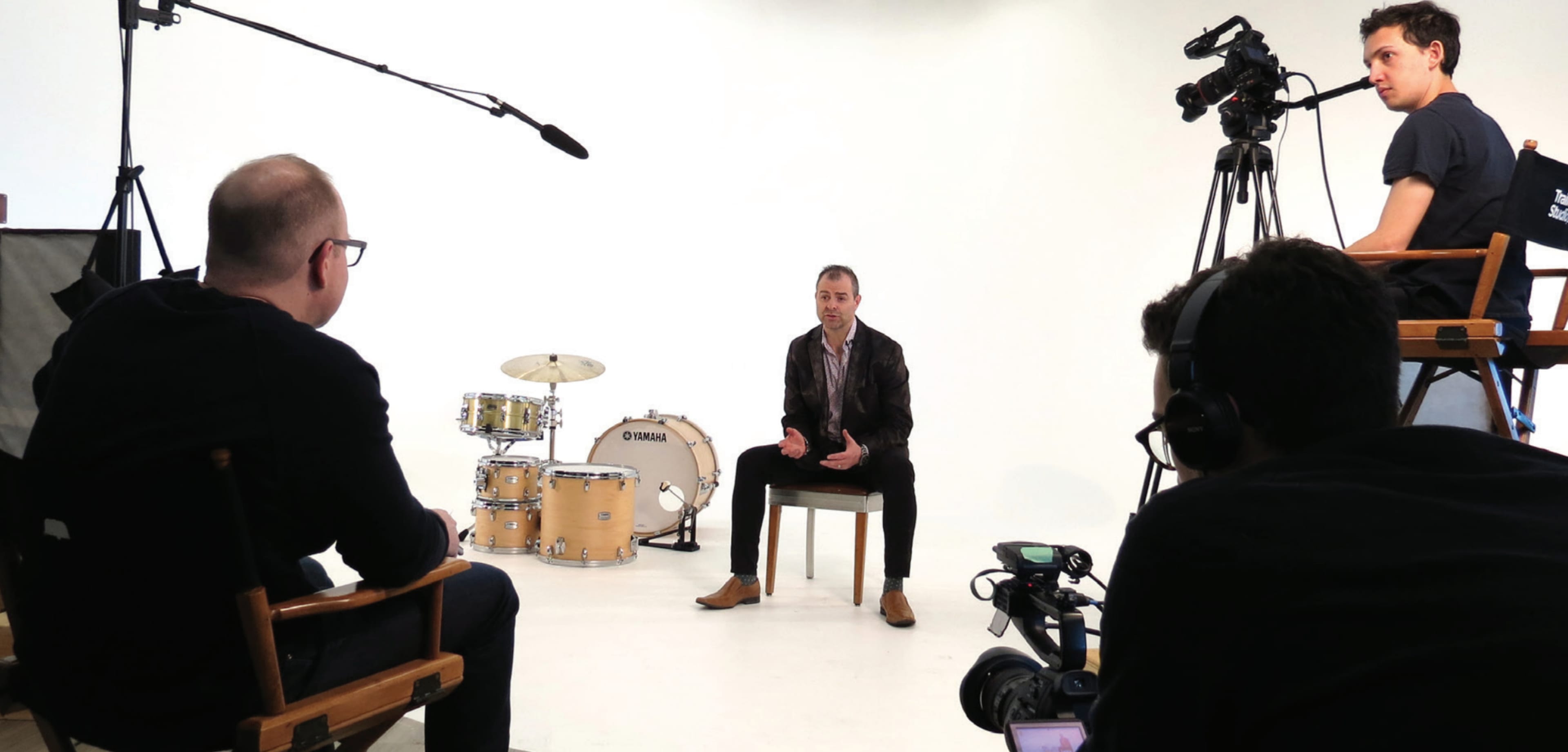 A man is seated on a chair in front of a drum set while being filmed by a crew with multiple cameras and lights in a studio setting.
