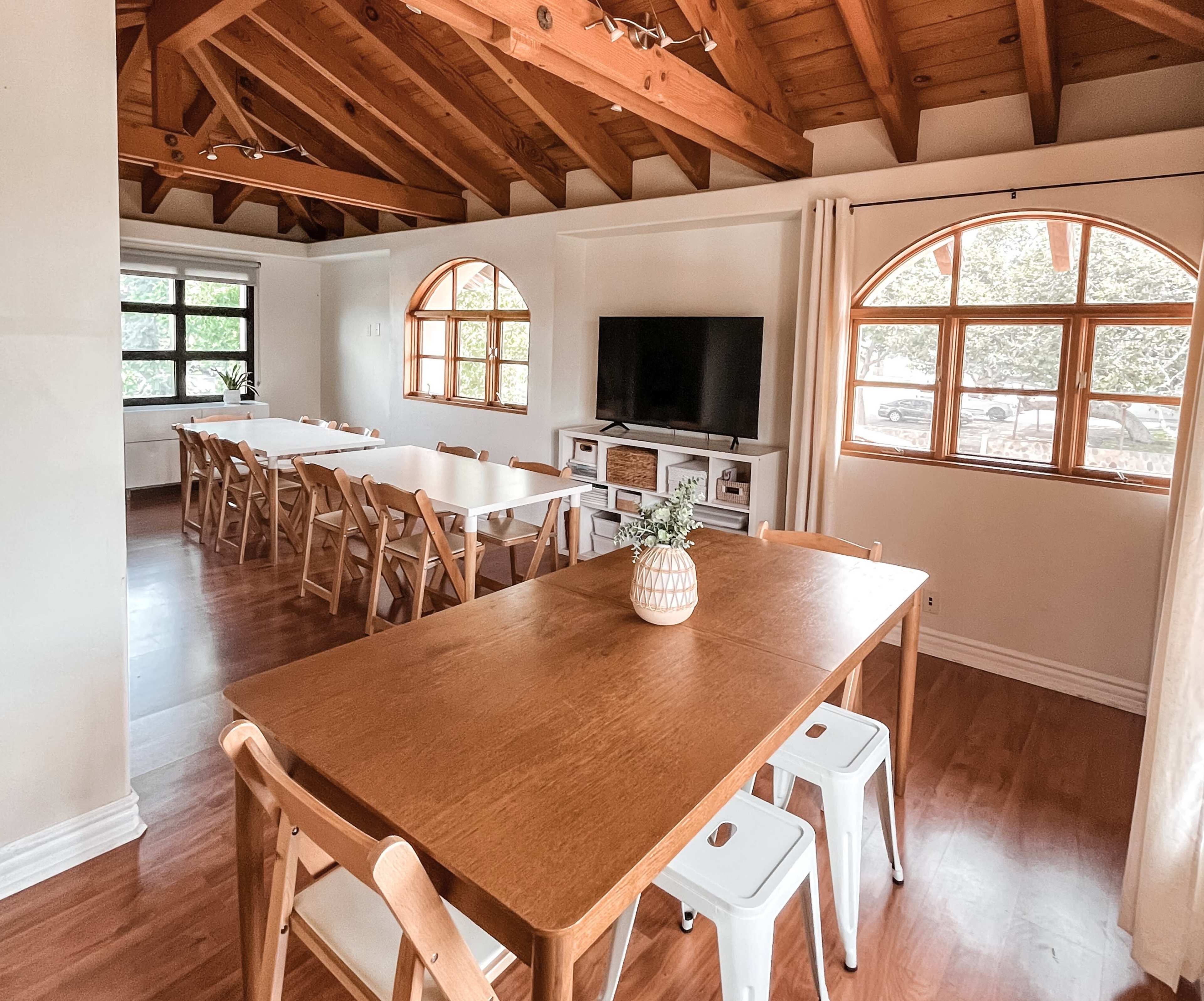 The image shows a spacious interior with wooden beams, two large dining tables, and a television against a wall, illuminated by natural light through arched windows.