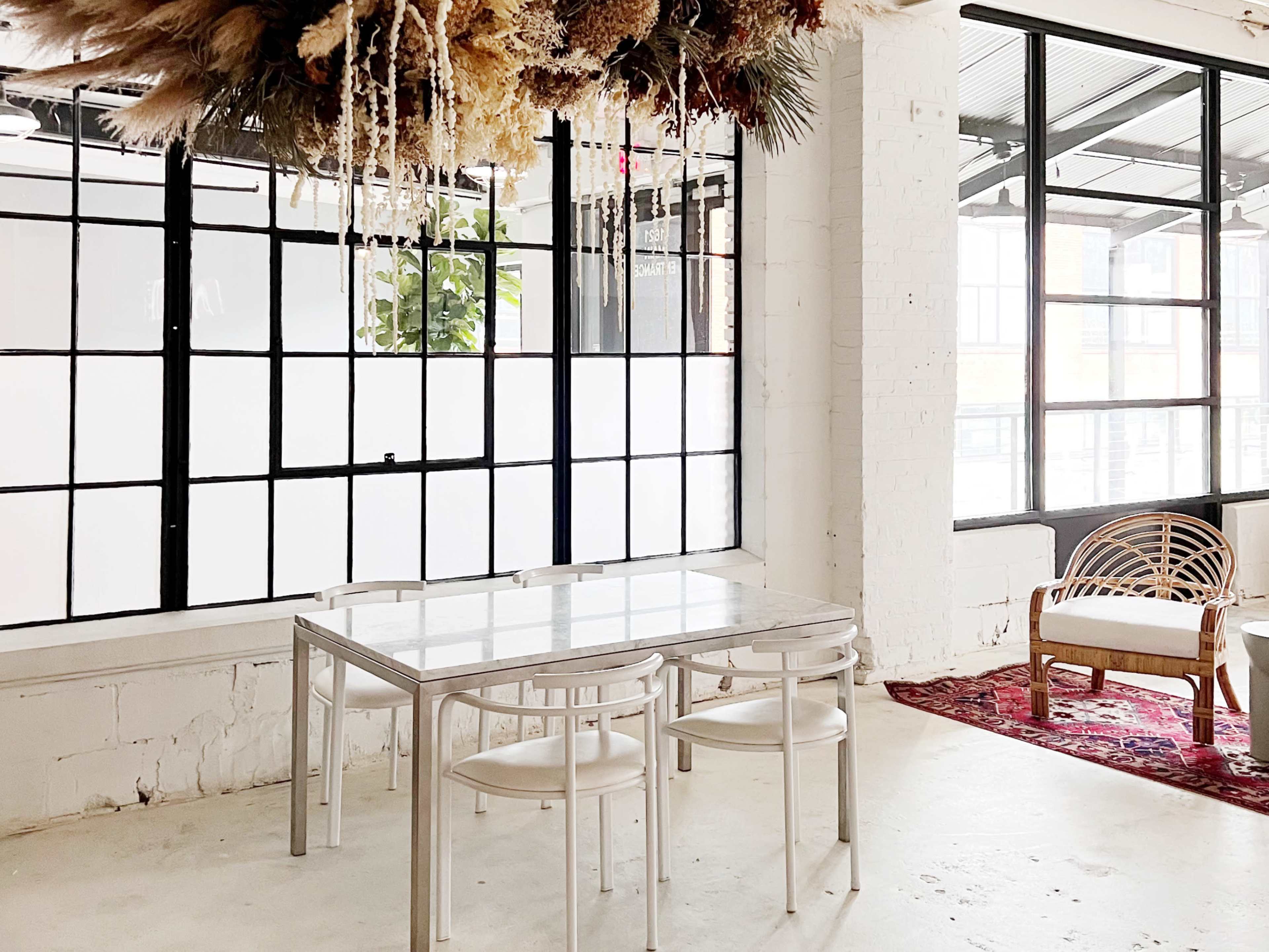 The image shows a bright, minimalist room with a glass table and three chairs next to a large window and a rattan chair on a patterned rug.