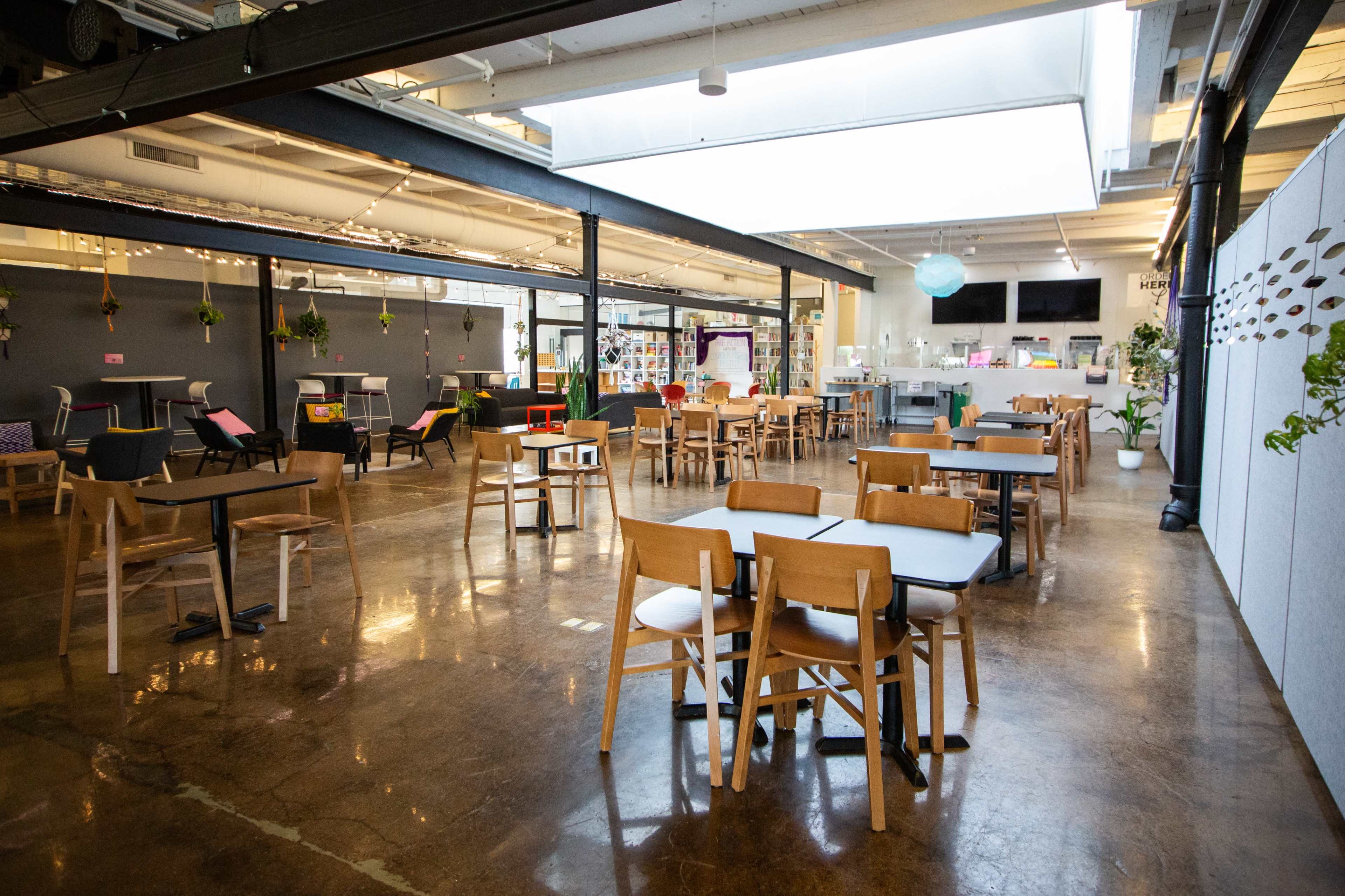 The image shows a spacious, modern workspace with several wooden tables and chairs arranged around a polished concrete floor, illuminated by natural light from overhead windows.