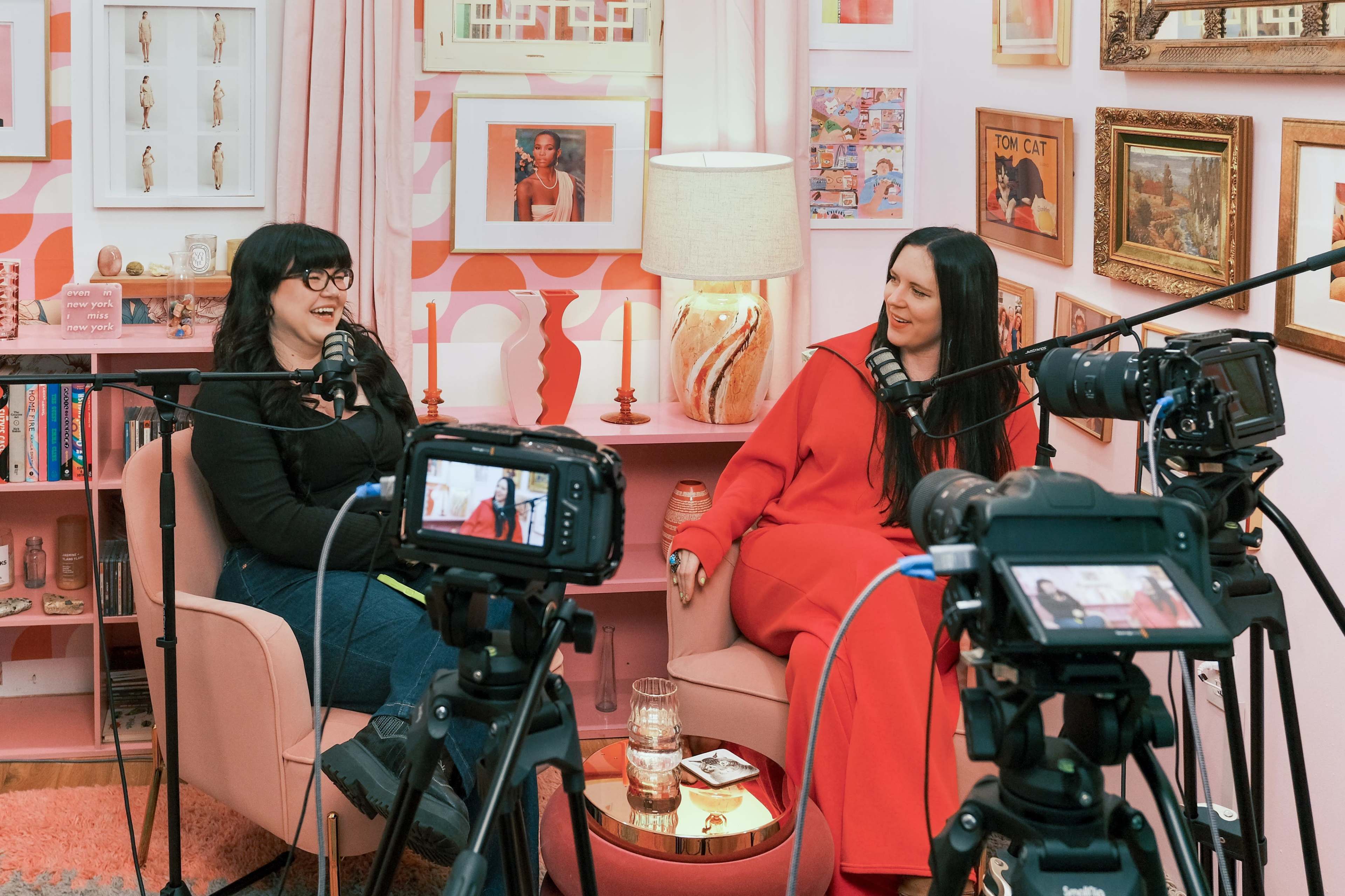 Two women sit in pink chairs in a brightly decorated room, with cameras positioned in front of them, suggesting a recording or interview setting.