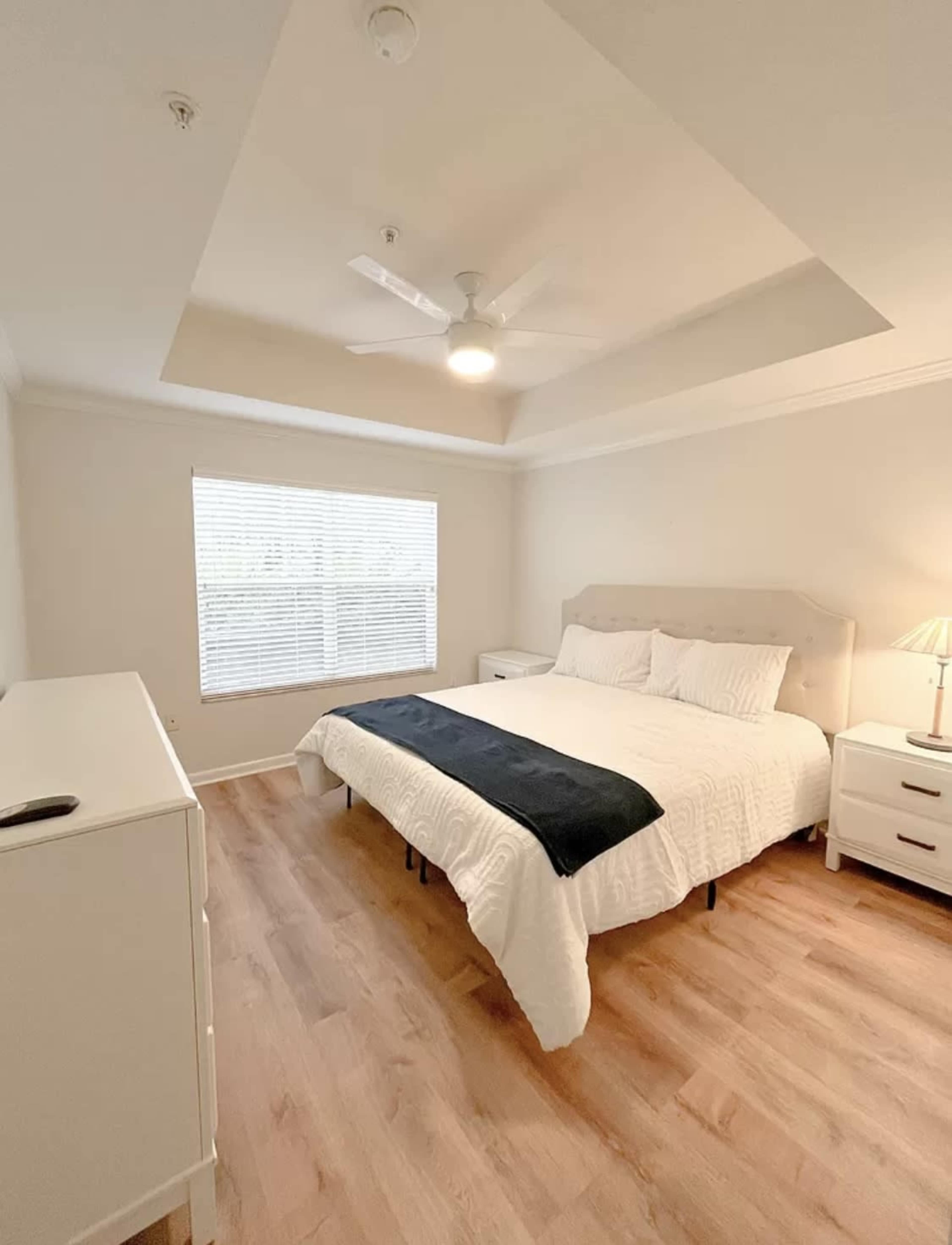 The image shows a neatly arranged bedroom featuring a large bed, a dresser, and a window with blinds.