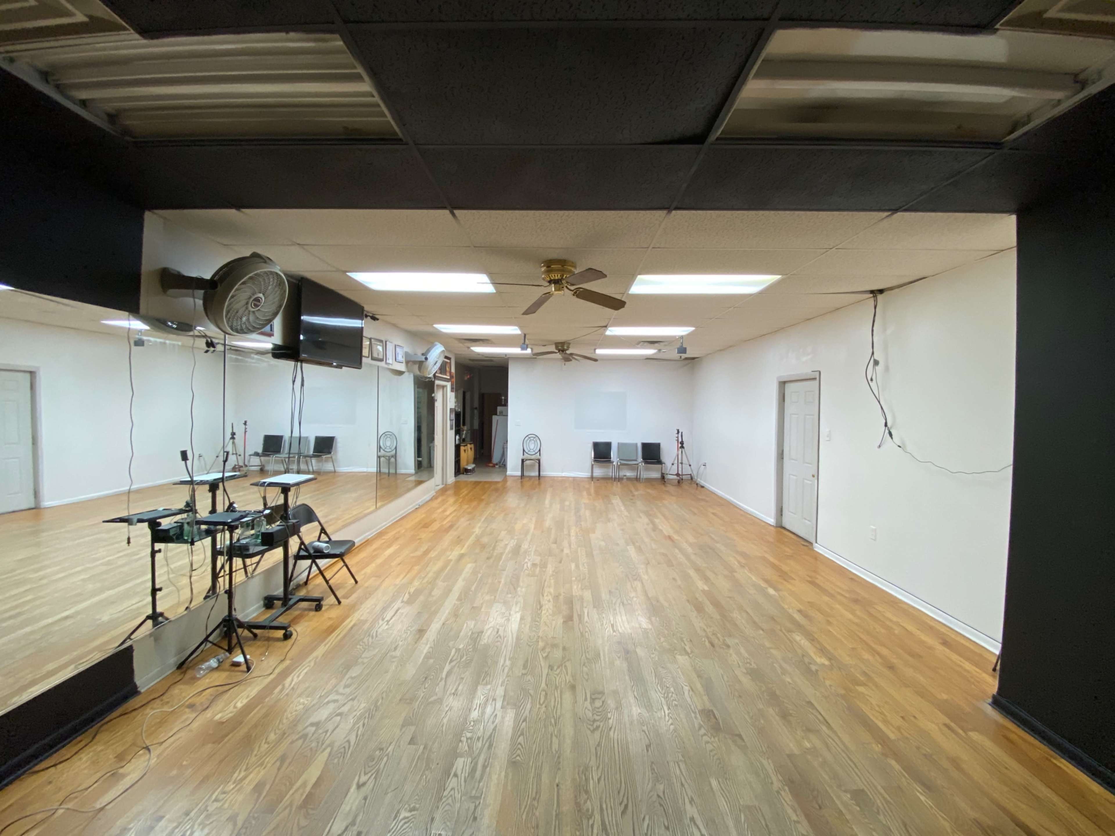 An empty room with hardwood floors, a ceiling fan, and mirrors along one wall, featuring a few chairs arranged in the corner.