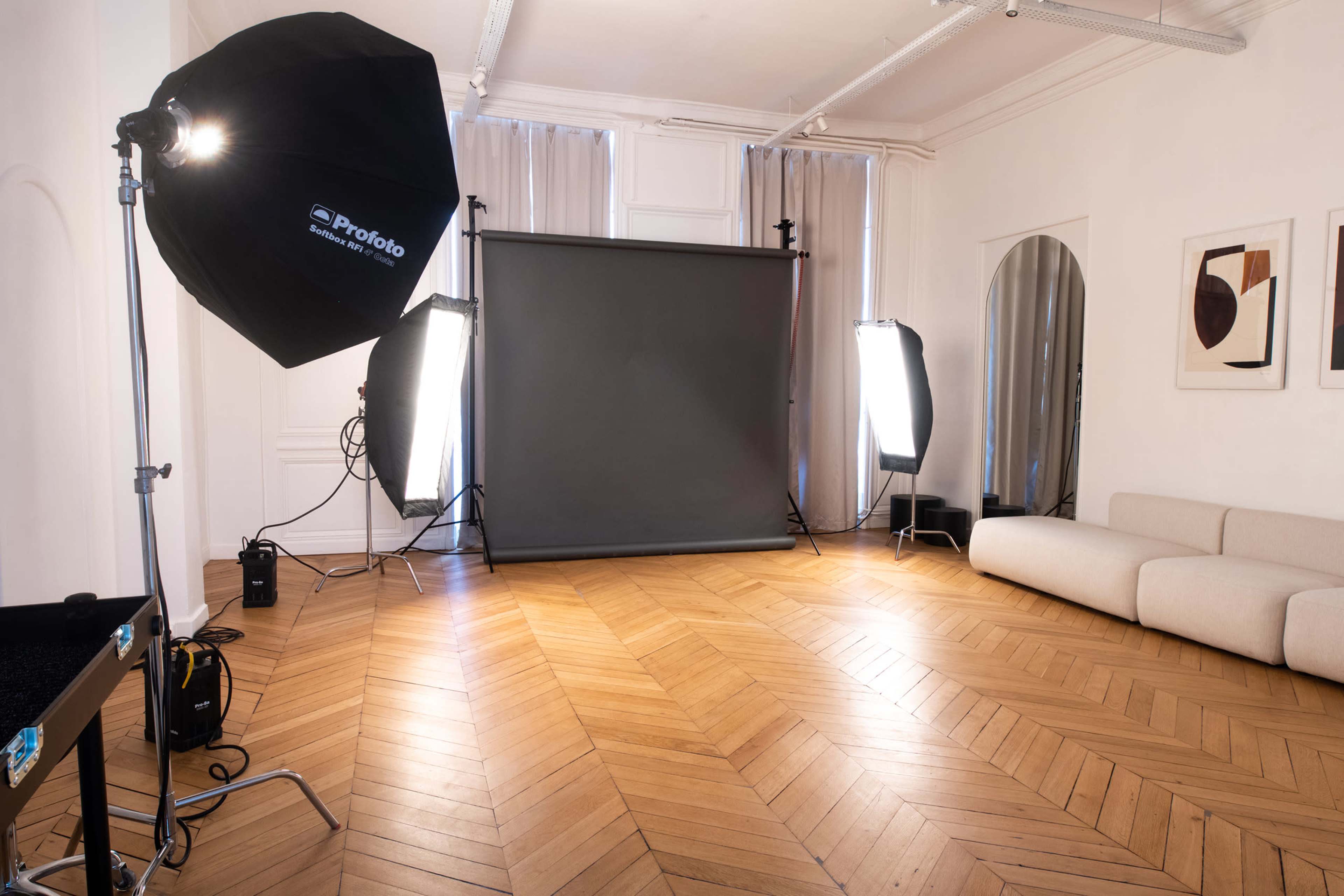 A photography studio with a gray backdrop, lighting equipment, and a light-colored sofa in a room featuring hardwood flooring.