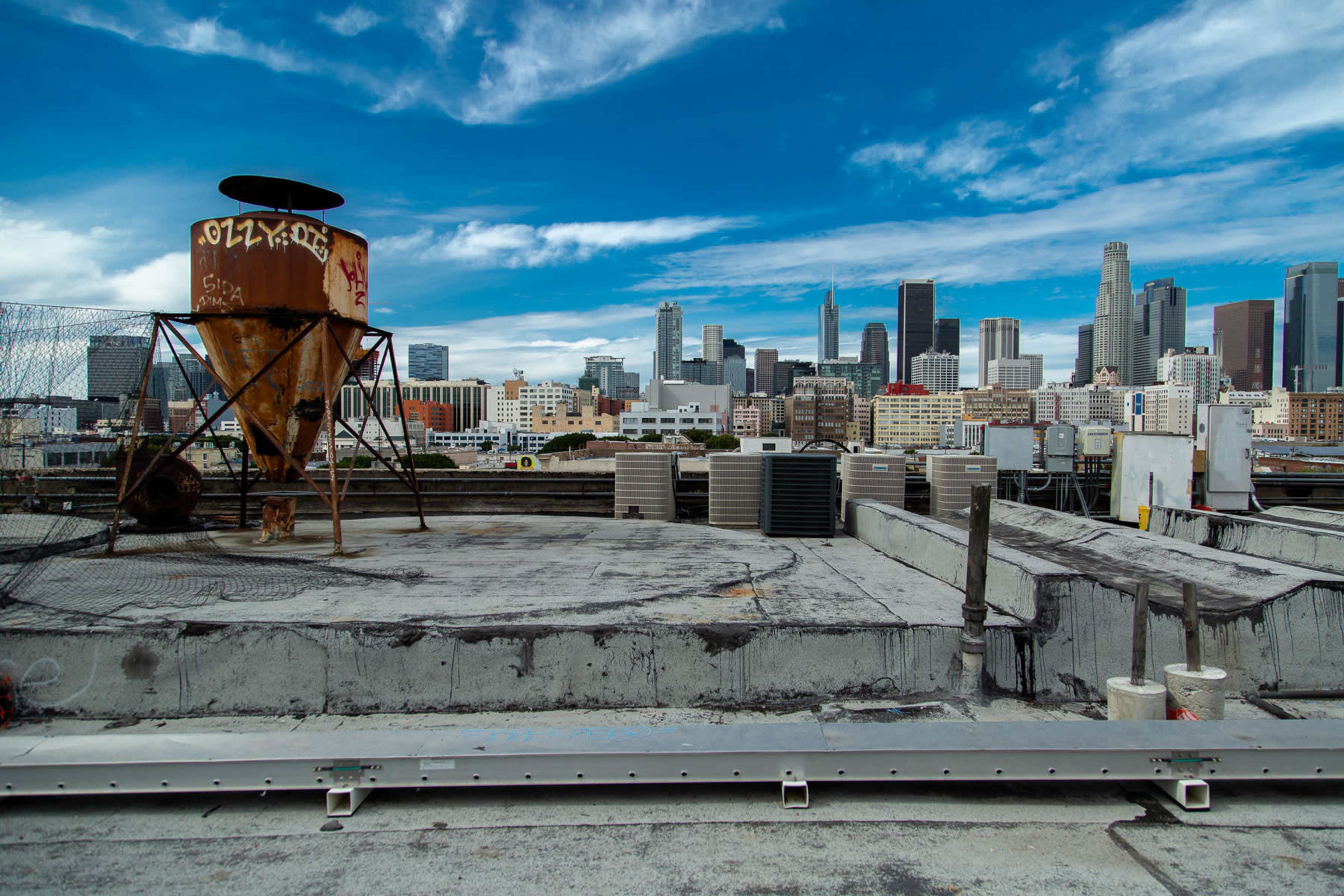 The image shows a rooftop view of a city skyline with a rusted container and various rooftop equipment in the foreground.