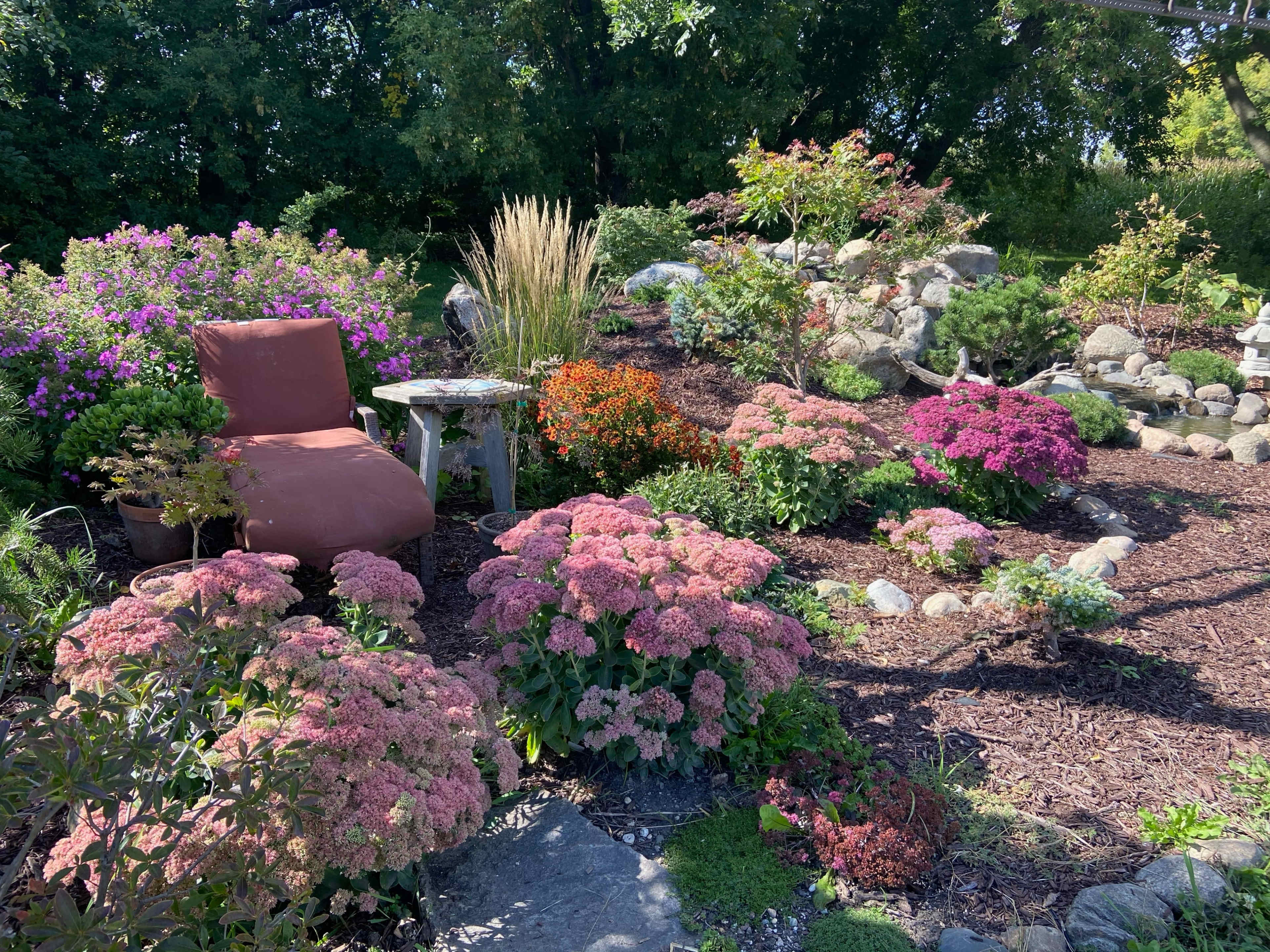 A cozy garden scene features a large chair amidst vibrant flowers and decorative rocks, with a small table nearby.